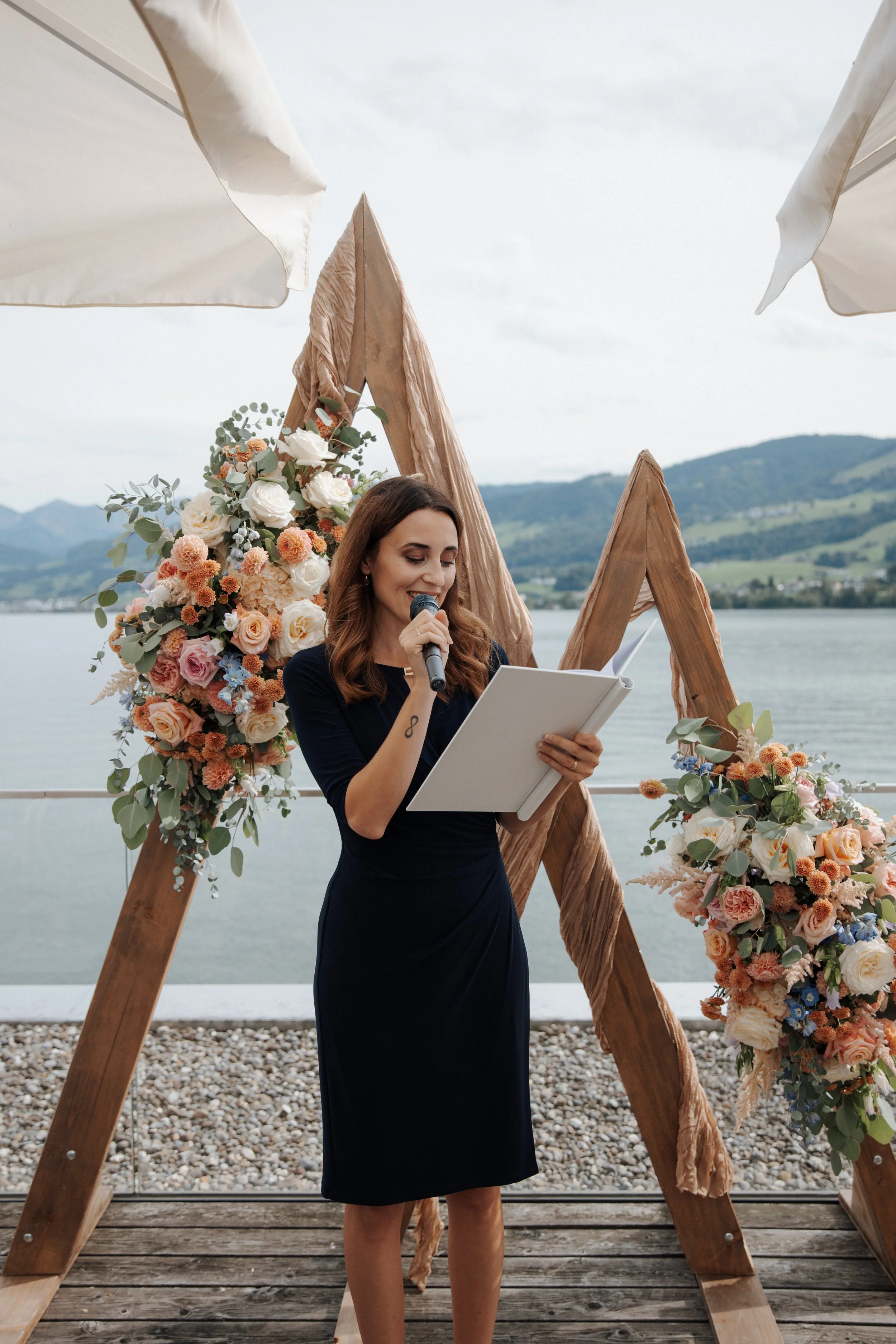 Frau liest bei einer Hochzeitszeremonie vor einem dekorierten Holzkreis am Wasser.