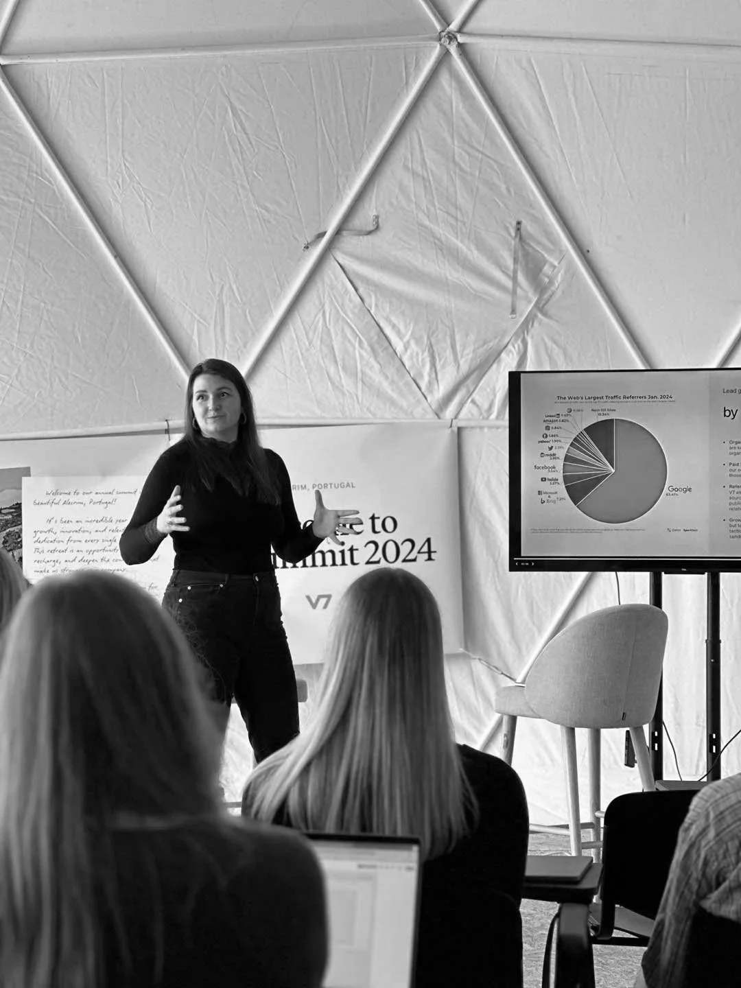 Our founder Veronika Vebere standing in front of an audience, giving a presentation at The V7 company summit in 202, with a large screen displaying a pie chart titled 'The Web's Largest Traffic Referrers Jan'