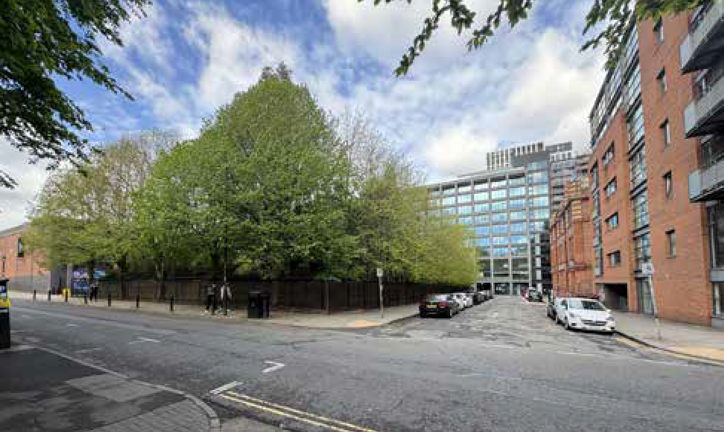City street corner with a large green tree, cars parked along the street, and modern brick and glass buildings under a partly cloudy sky.