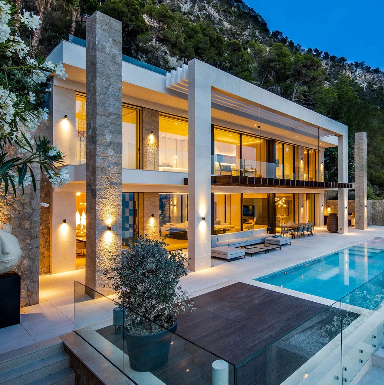 Modern, luxurious two-story house with large glass windows, outdoor pool, and seating area, set against a hillside with trees at dusk.