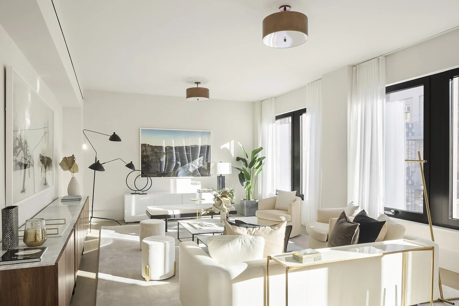 Bright and modern living room with white walls, large windows with white curtains, beige armchairs, a white sofa, a black coffee table, a tall green plant, and minimalist decor, including a framed landscape photograph on the wall.