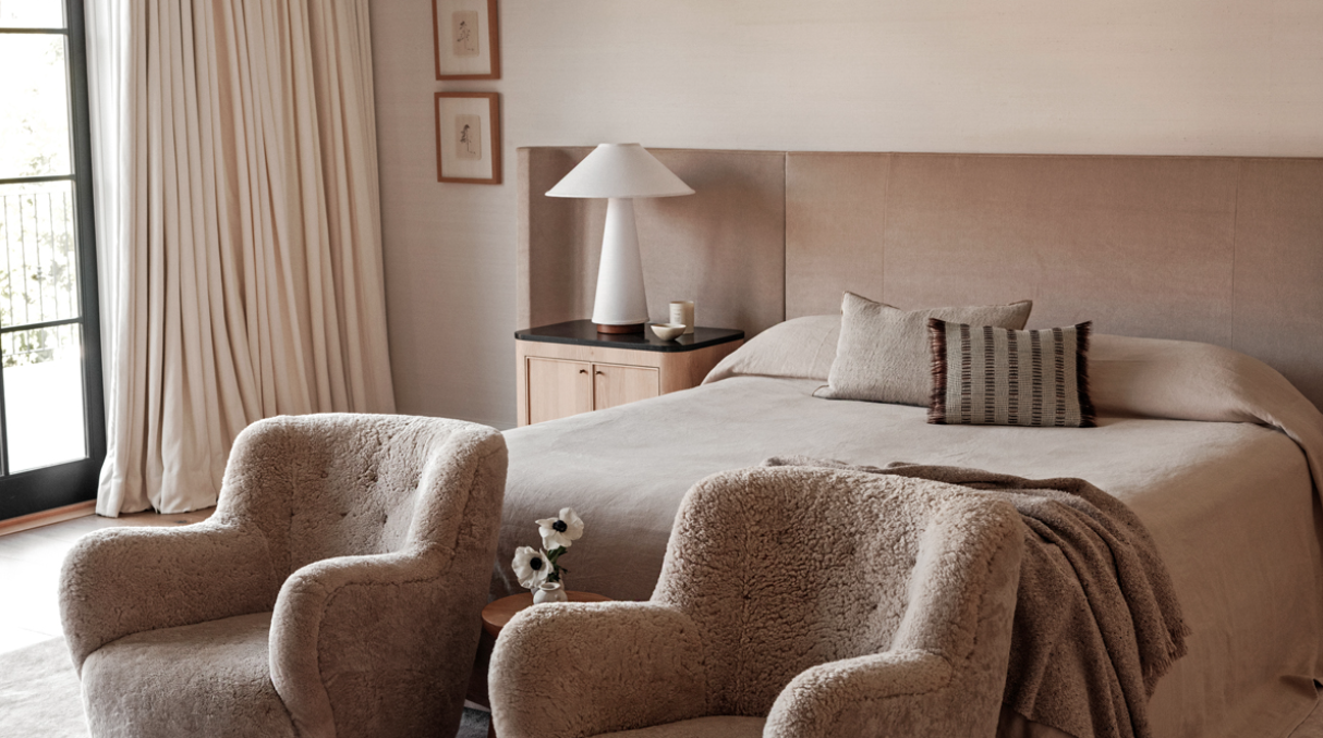 A cozy bedroom with a bed, two plush armchairs, a nightstand with a lamp, and a sliding glass door with long curtains.