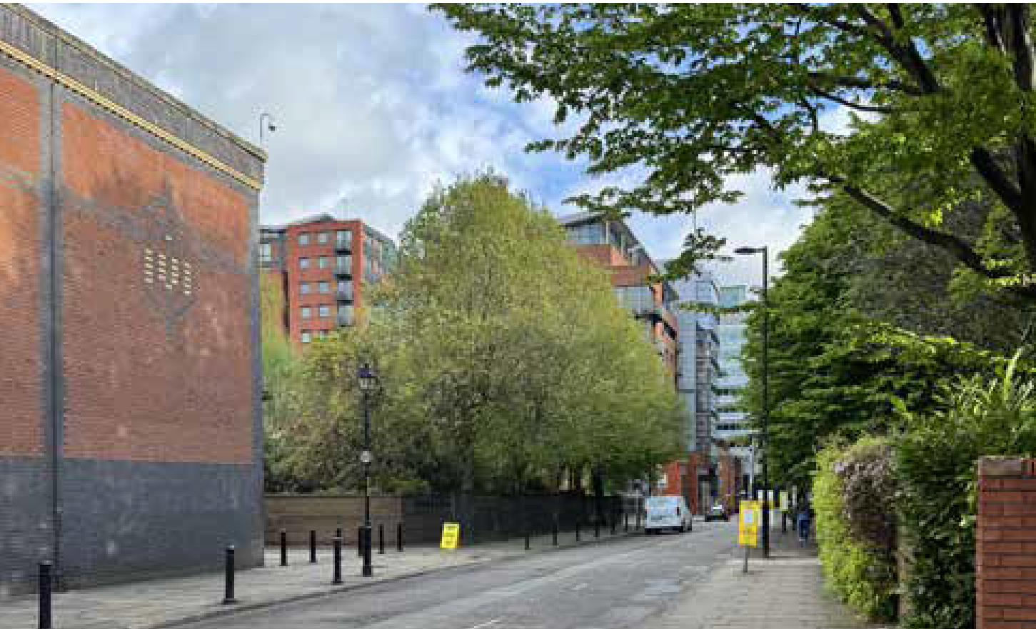 A city street scene with trees, buildings, a lamppost, a white van, and a few pedestrians on a partly cloudy day.