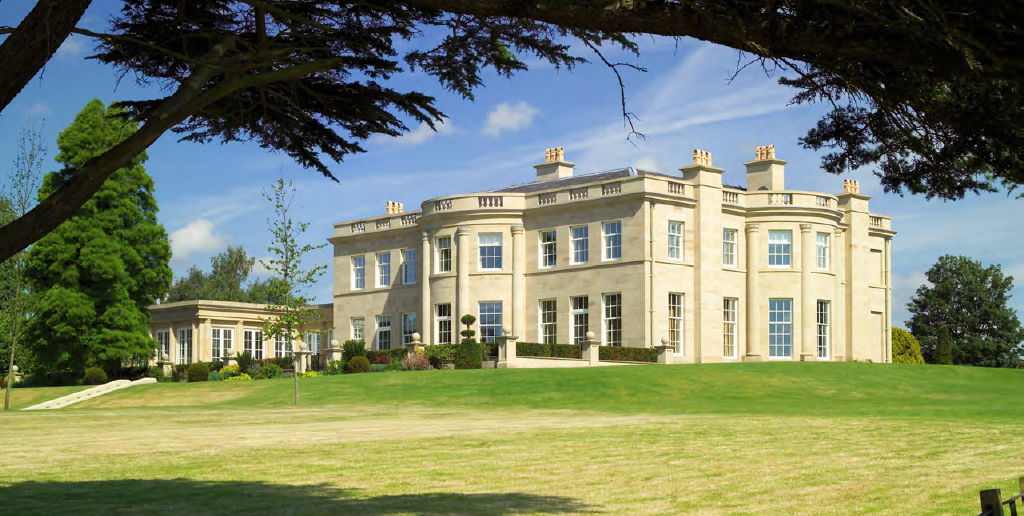A large, elegant mansion with a light-colored facade and numerous windows, surrounded by lush green lawn and trees under a partly cloudy sky.