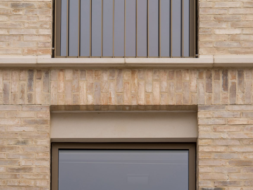 Close-up of a brick building with a window and balcony, showing beige bricks, a window with a metal frame, and a metal balcony railing.