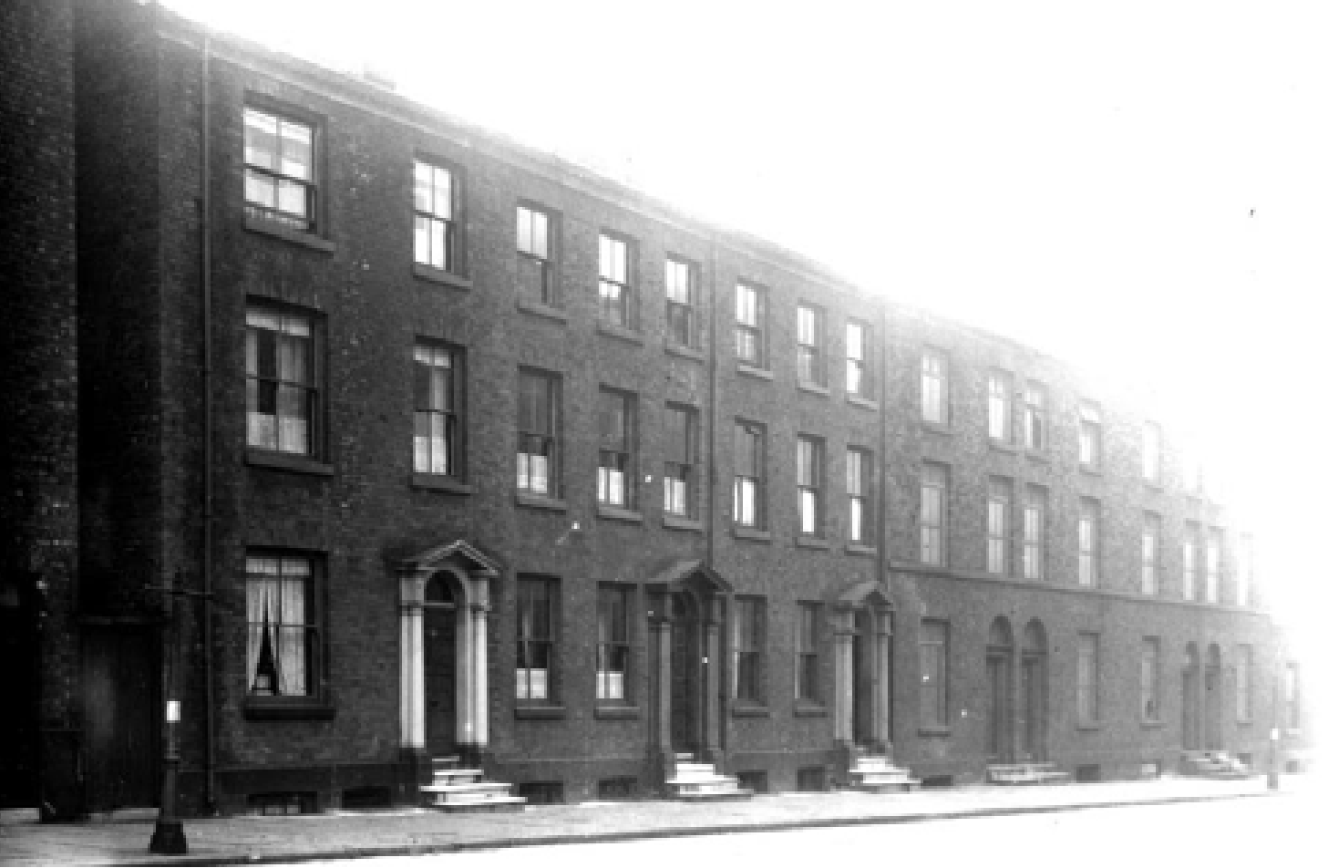 Black and white photo of a row of brick apartment buildings with steps leading up to front doors.