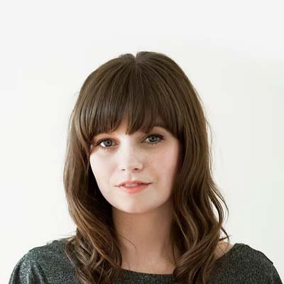 Portrait of a young woman with shoulder-length brown hair and bangs, wearing a dark top, against a light background.