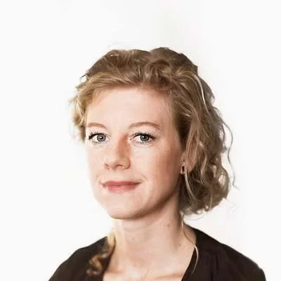 A woman with curly blonde hair and blue eyes, wearing a black top, looking at the camera with a slight smile.