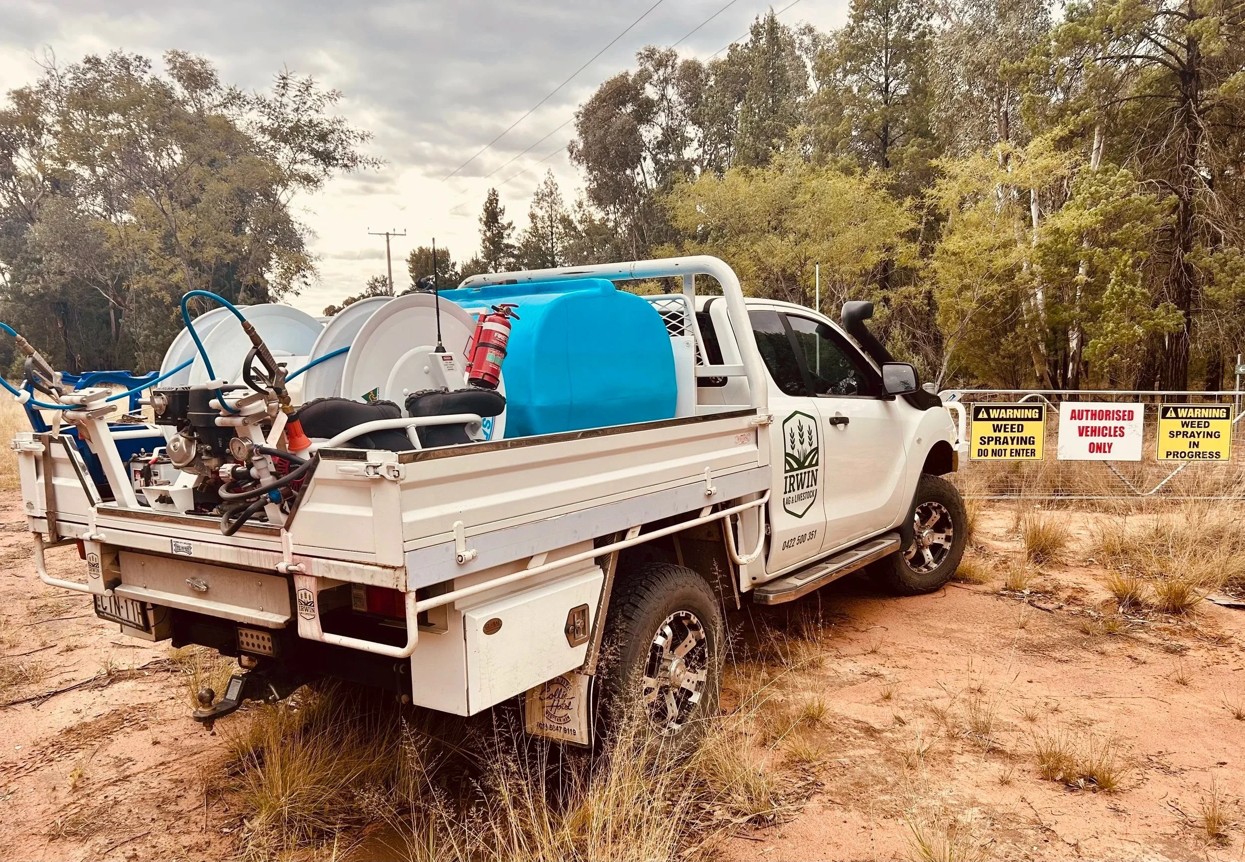 A white utility truck carrying spraying equipment, including hoses, a blue tank, and a fire extinguisher, parked outdoors near warning signs about weed spraying in progress, with trees and cloudy sky in the background.