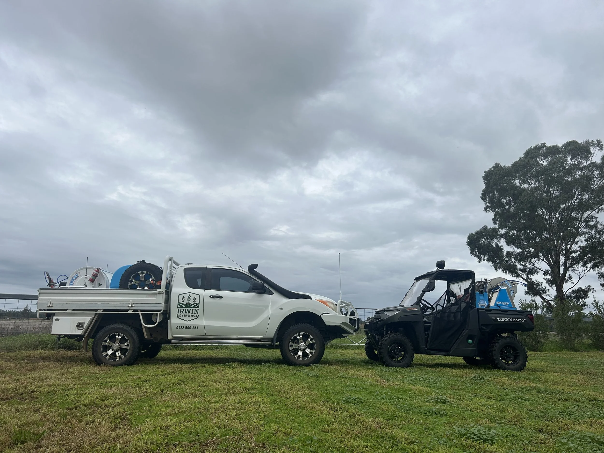 A white pickup truck with a logo for IRWIN A&G & LIVESTOCK and a phone number, parked next to an all-terrain vehicle, both on a grassy field. The truck bed has equipment, and there are trees and cloudy skies in the background.