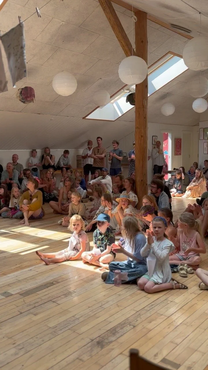 Billede af en gruppe børn og voksne, der sidder og står i et lyst rum med træloft og store spænder, og ser til en begivenhed. Der er papirlamper og store ovenlys, der giver naturligt lys.