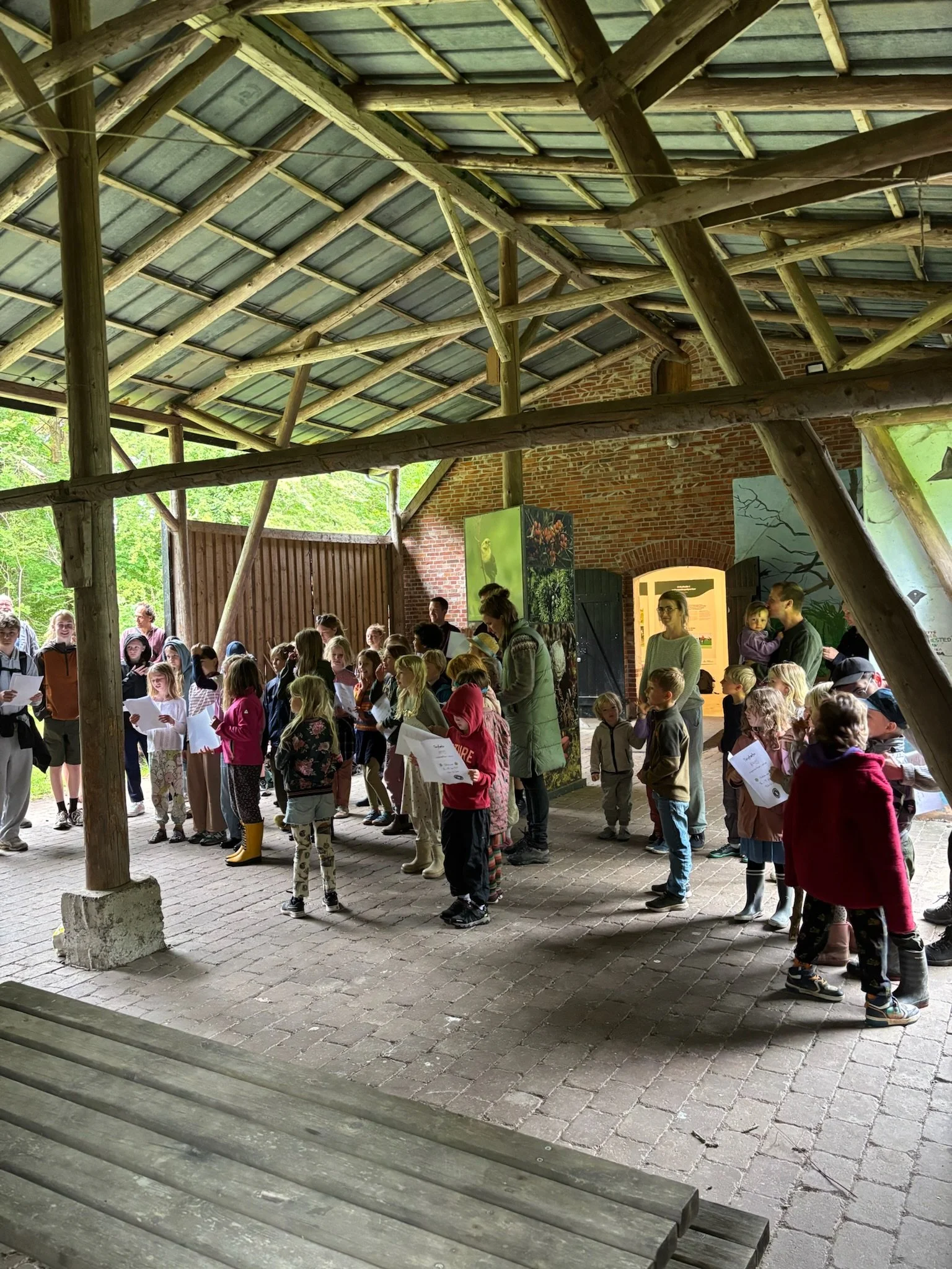 Børnegruppe samlet under et tag i et udendørs område, der er bygget med træstolper og tage, med nogle voksne tilstede, i en naturkulisse.