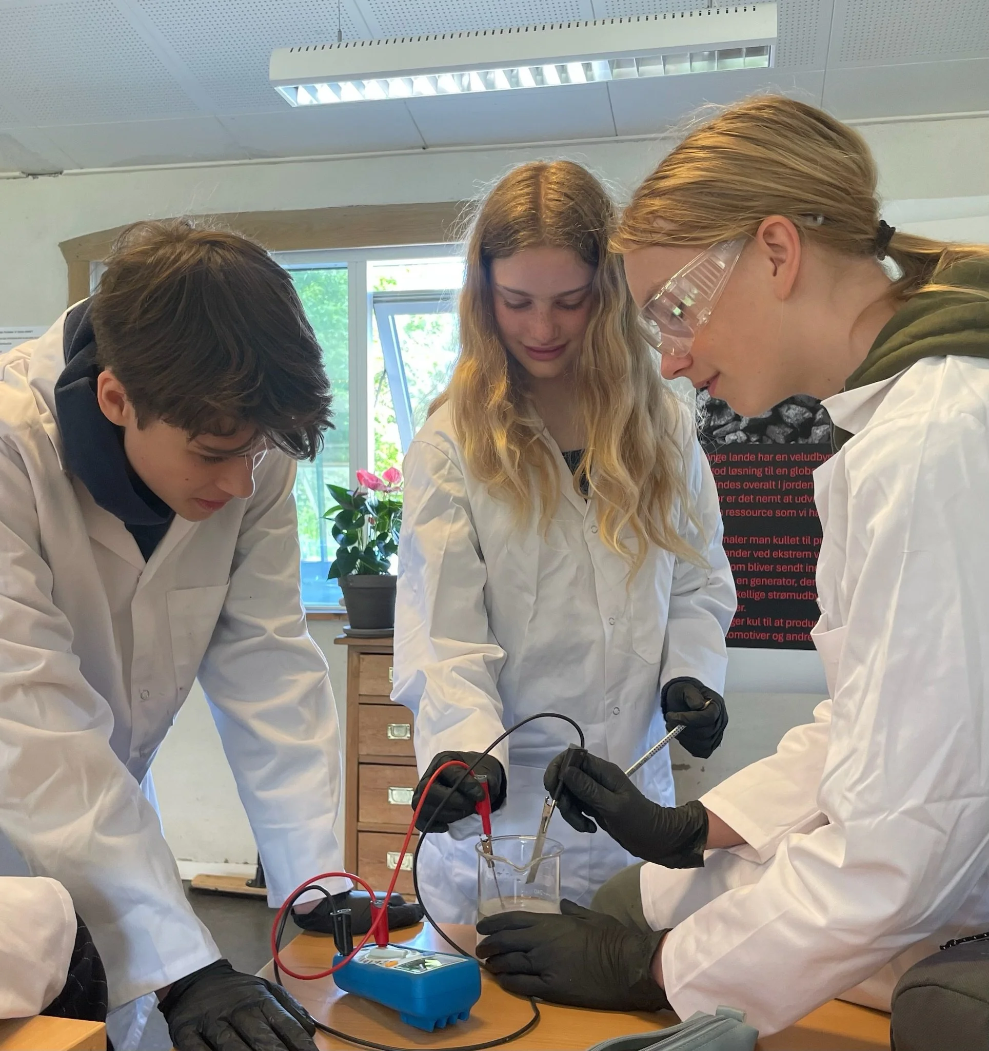 Tre unge voksne i laboratoriefrakker arbejder med laboratorieudstyr, herunder en elektrokemisk måling, i et laboratoriemiljø.