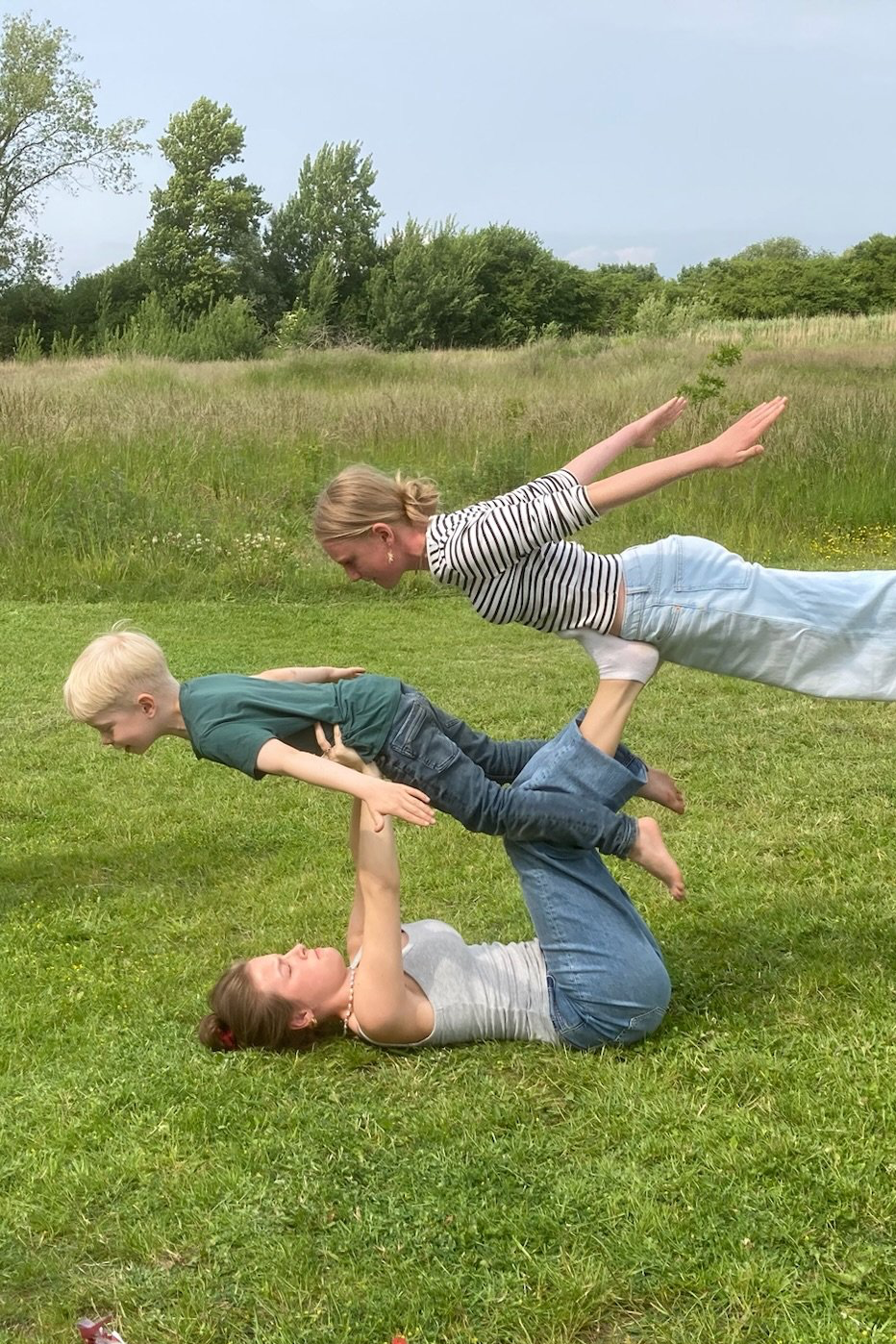 Tre personer laver en menneskelig pyramide foran et grønt landskab med træer i baggrunden.