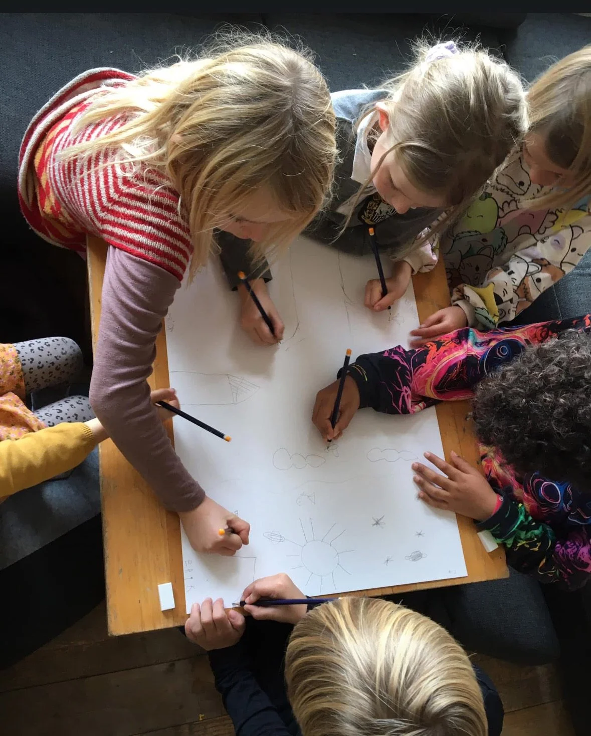 Børn og en voksen tegner på et stort stykke papir på et bord, tegner blandt andet en sol.