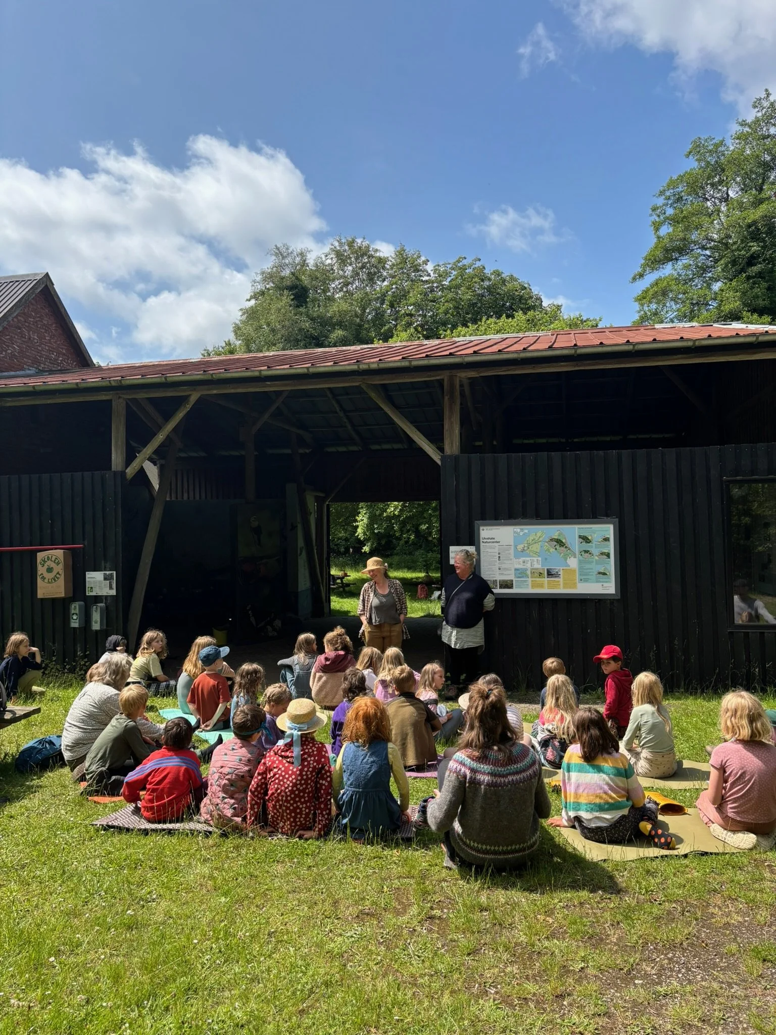 En gruppe børn sidder på måtter foran en udendørs informationsskærm, mens to voksne står foran dem under et træpergola, i en grøn park med træer under en blå himmel med hvide skyer.