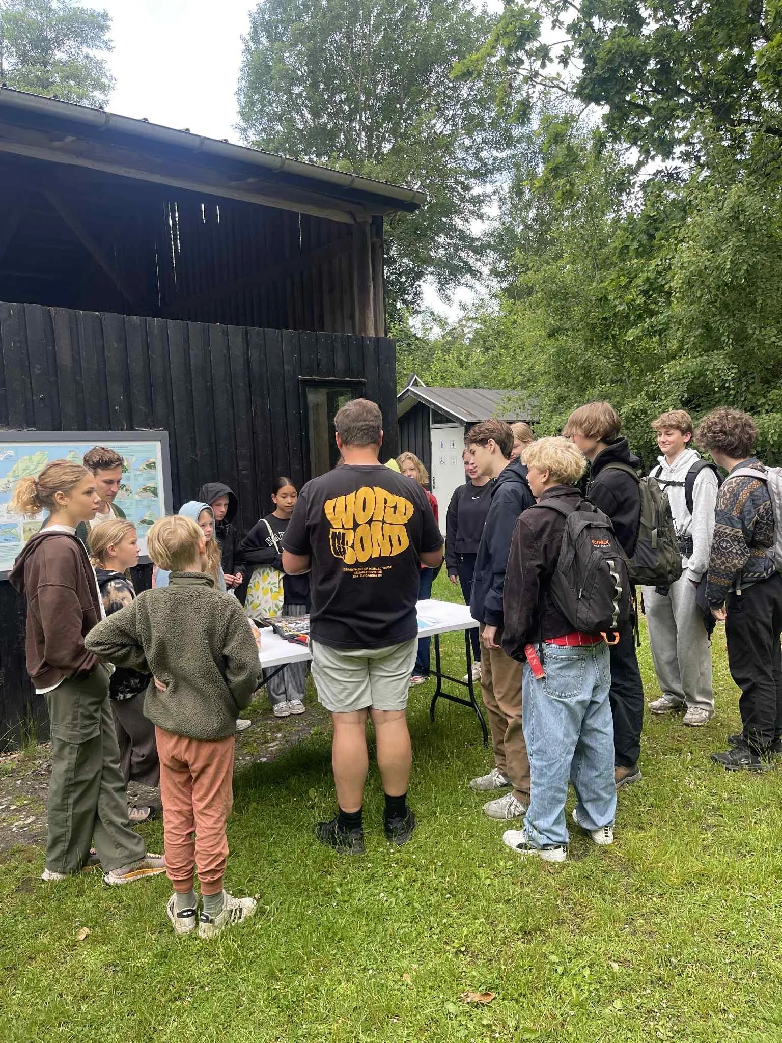 En gruppe unge mennesker står omkring en bord, hvor en instruktør giver en forklaring, uden for et grønt område med træer.