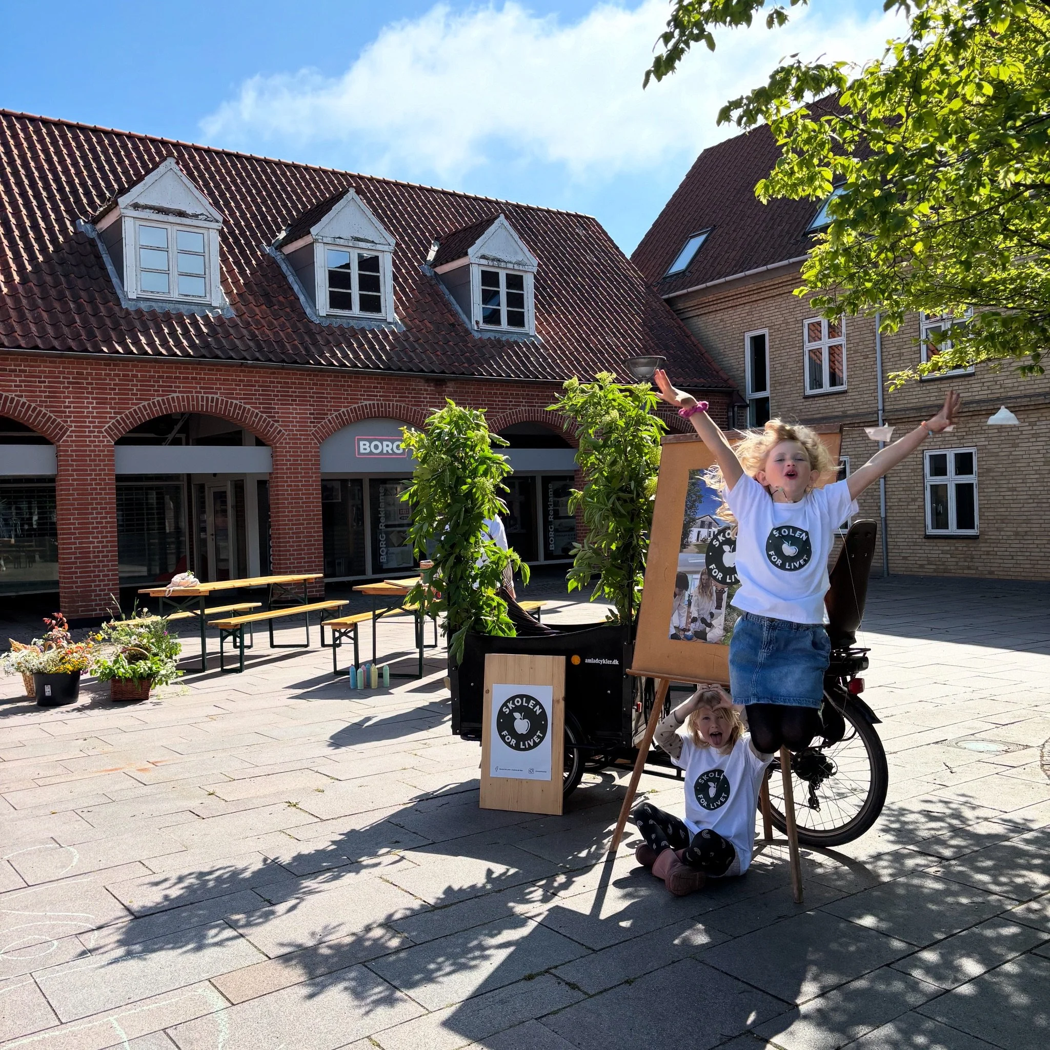 To unge piger med Hvide t-shirts og sort leggings leger udenfor på en byrundtur. Den ene pige sidder på jorden og den anden hopper op i luften. Der er planter og billed stel foran en cykel. Billedet er taget på en solrig dag i en by med gamle røde og gule murhuse.