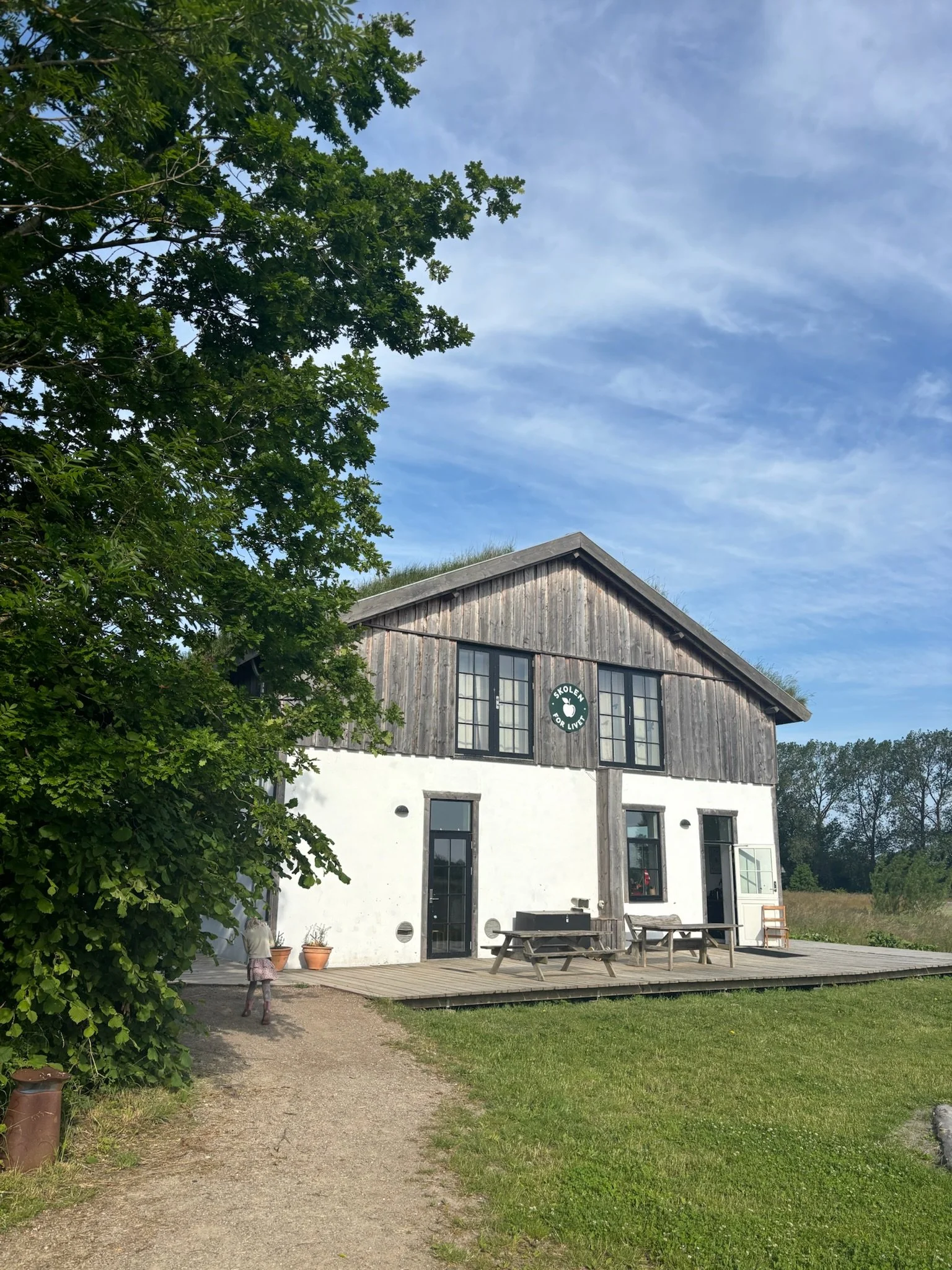 En bygning med træ- og hvide vægge, store vinduer, en træterrasse og en grøn græsplæne foran. Der er en pige, der går på en sti. Træer og en blå himmel med spredte skyer i baggrunden.