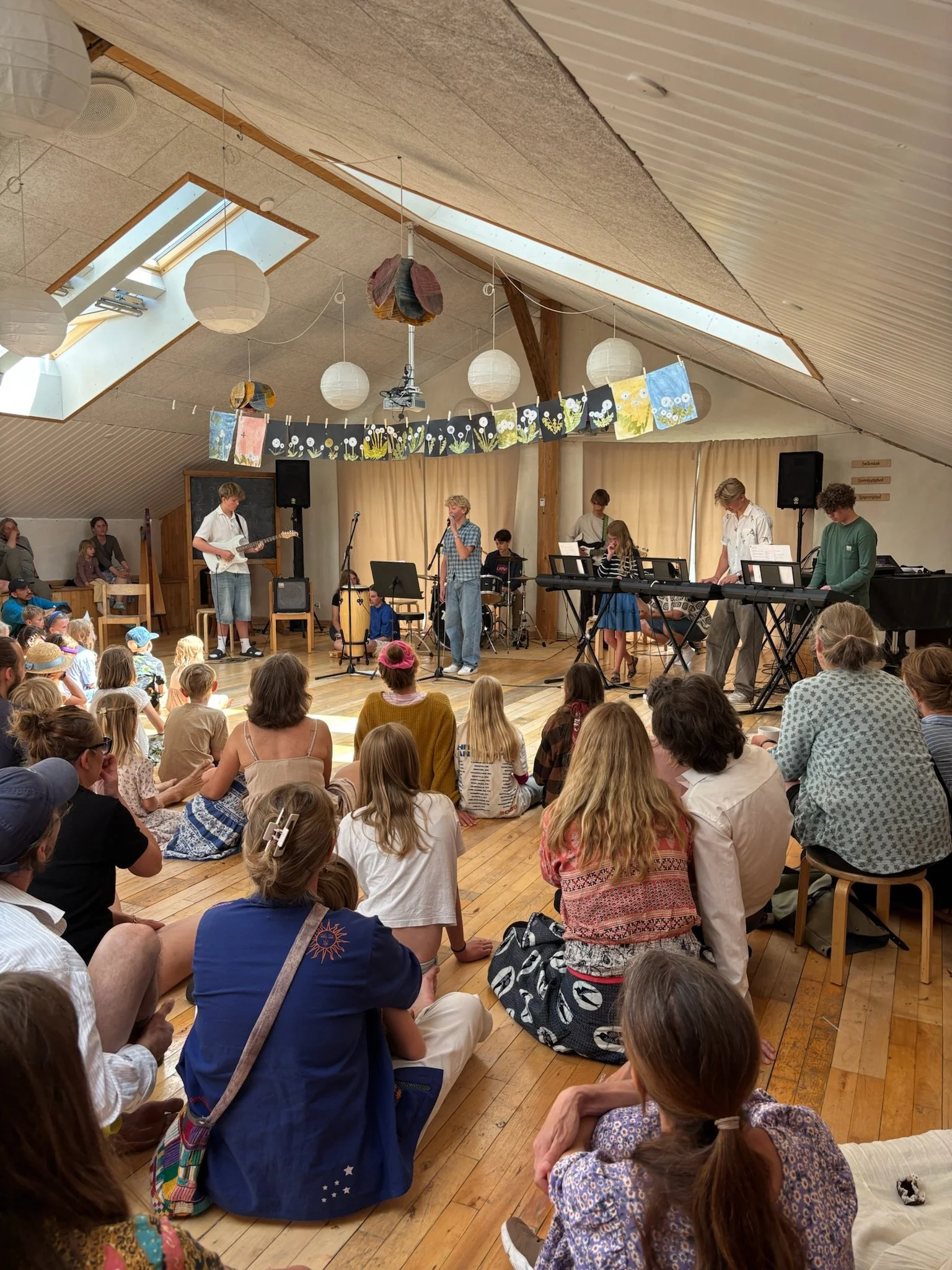 En koncert i et loftsrum med børn og voksne, hvor en musikgruppe spiller på scenen, omgivet af himmelvinduer og dekorationer i loftet.