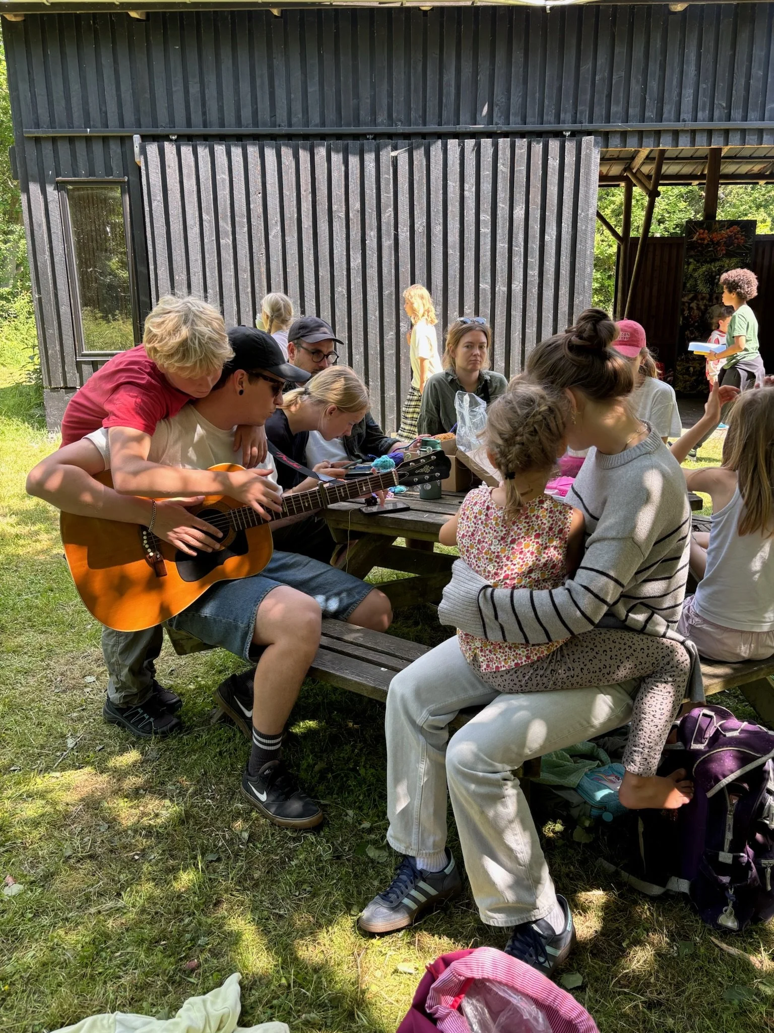 Børn og voksne sidder udenfor ved et udendørs picnicbord, hvor nogle spiller musik, bl.a. en der spiller guitar, og nogle andre hygger sig, mens nogle mennesker står og går i baggrunden.