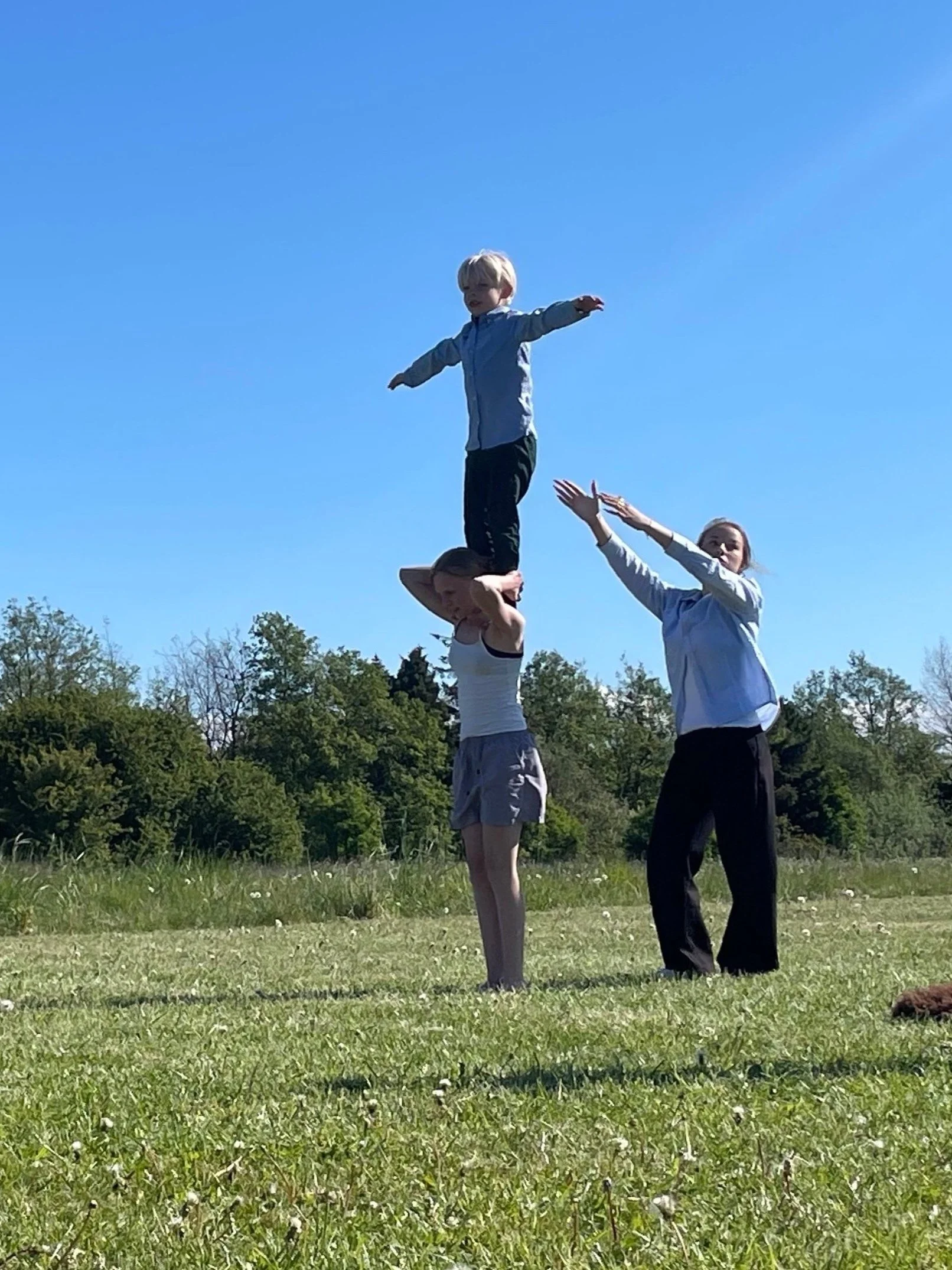 Tre børn laver en akrobatisk opstilling i en grøn park under en klar blå himmel, hvor den midterste pige holder en dreng oppe på skuldrene.