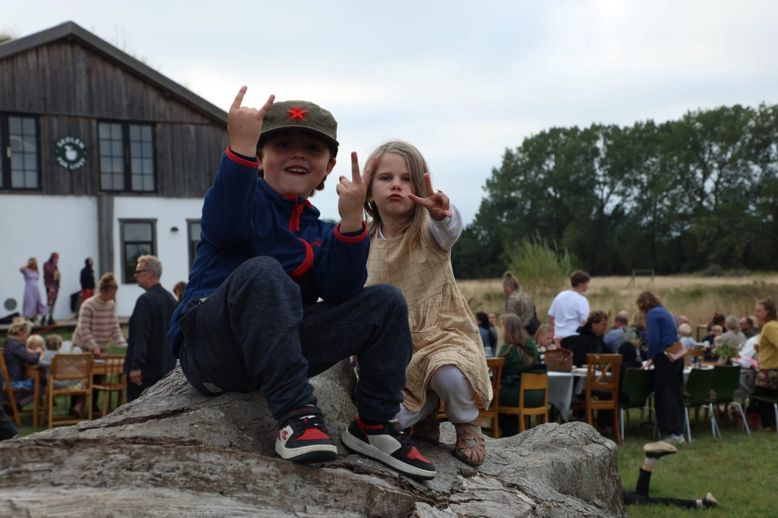 To børn sidder på en stor sten, de gør rock and roll-gestus med hænderne på en festival eller udendørs arrangement, med mange mennesker og et gammelt træhus i baggrunden.