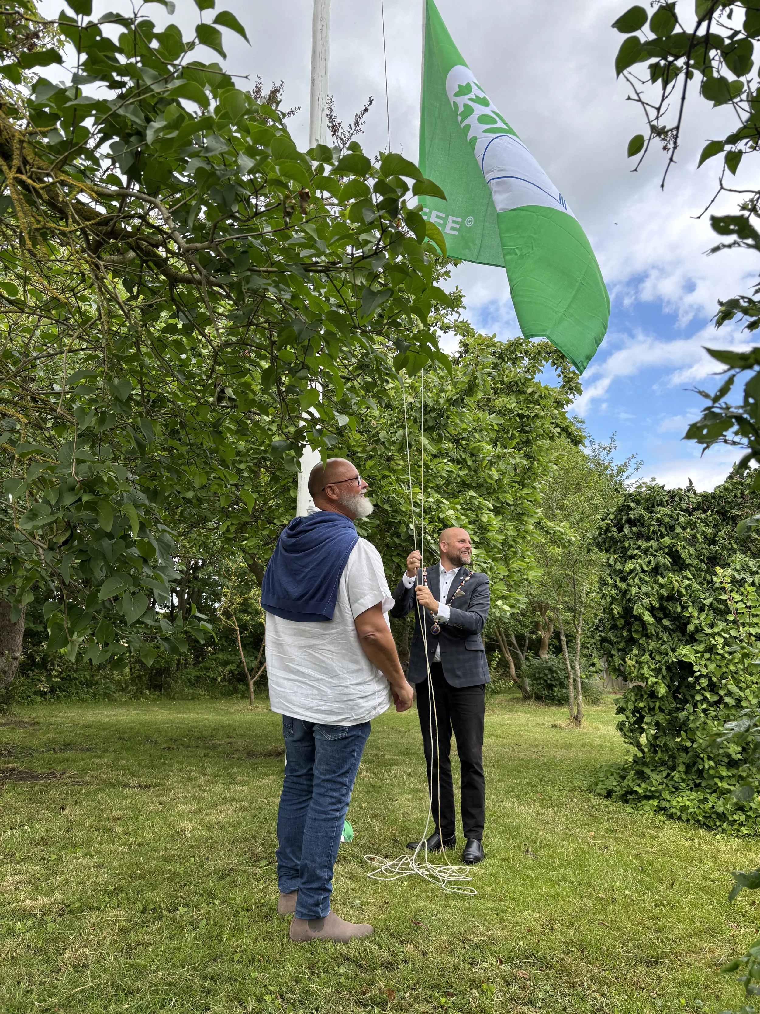 To mænd hænger en grøn protestflag i et træ i en have med grønne træer og græs. Den ene mand holder i flagets snor, mens den anden står ved siden og ser op på flaget.