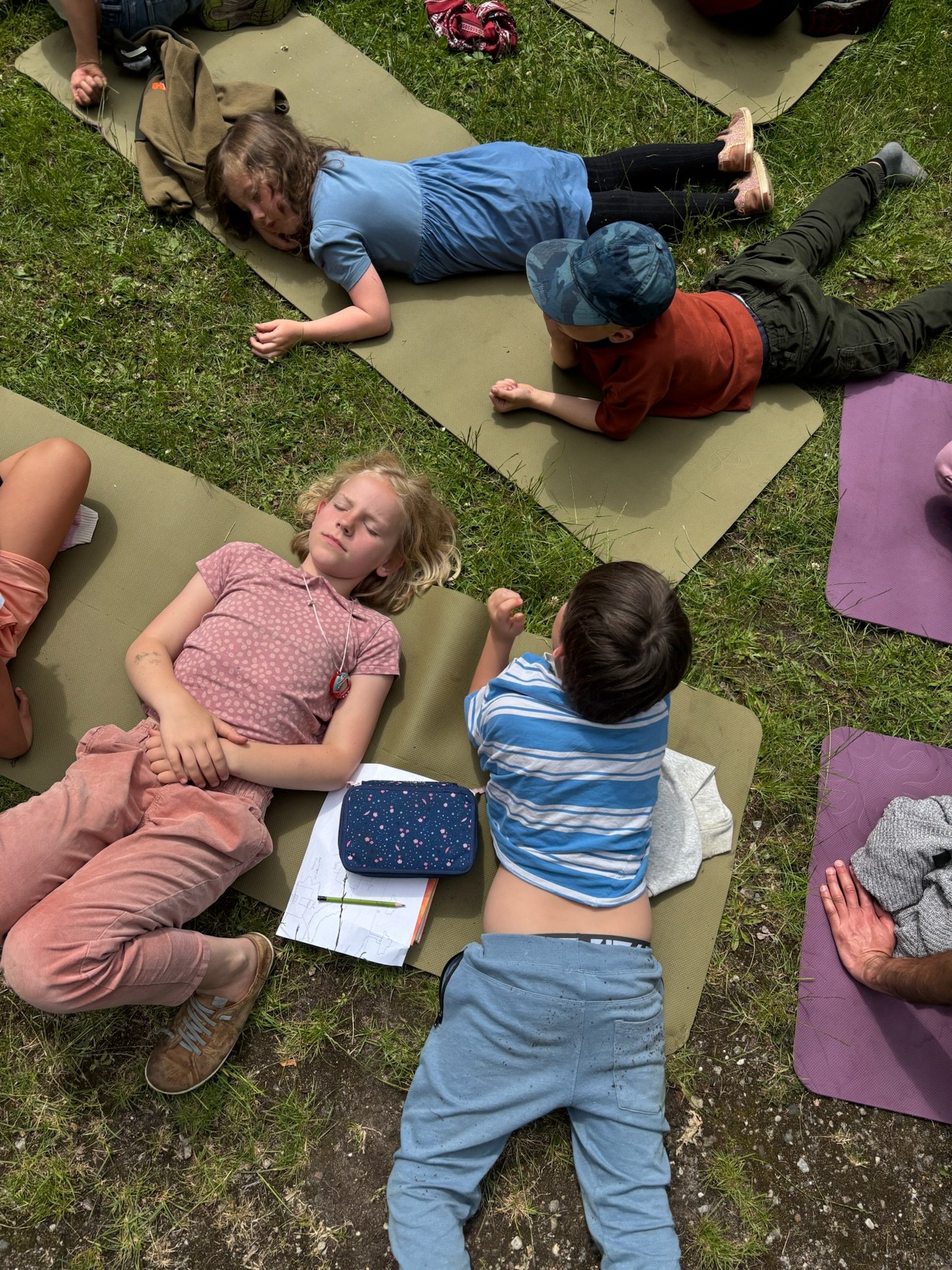 Børn ligger på yogamåtter på græs, nogle hviler, anden taler, og en pige med lommeur og tegning ved siden af ser ud til at hvile.
