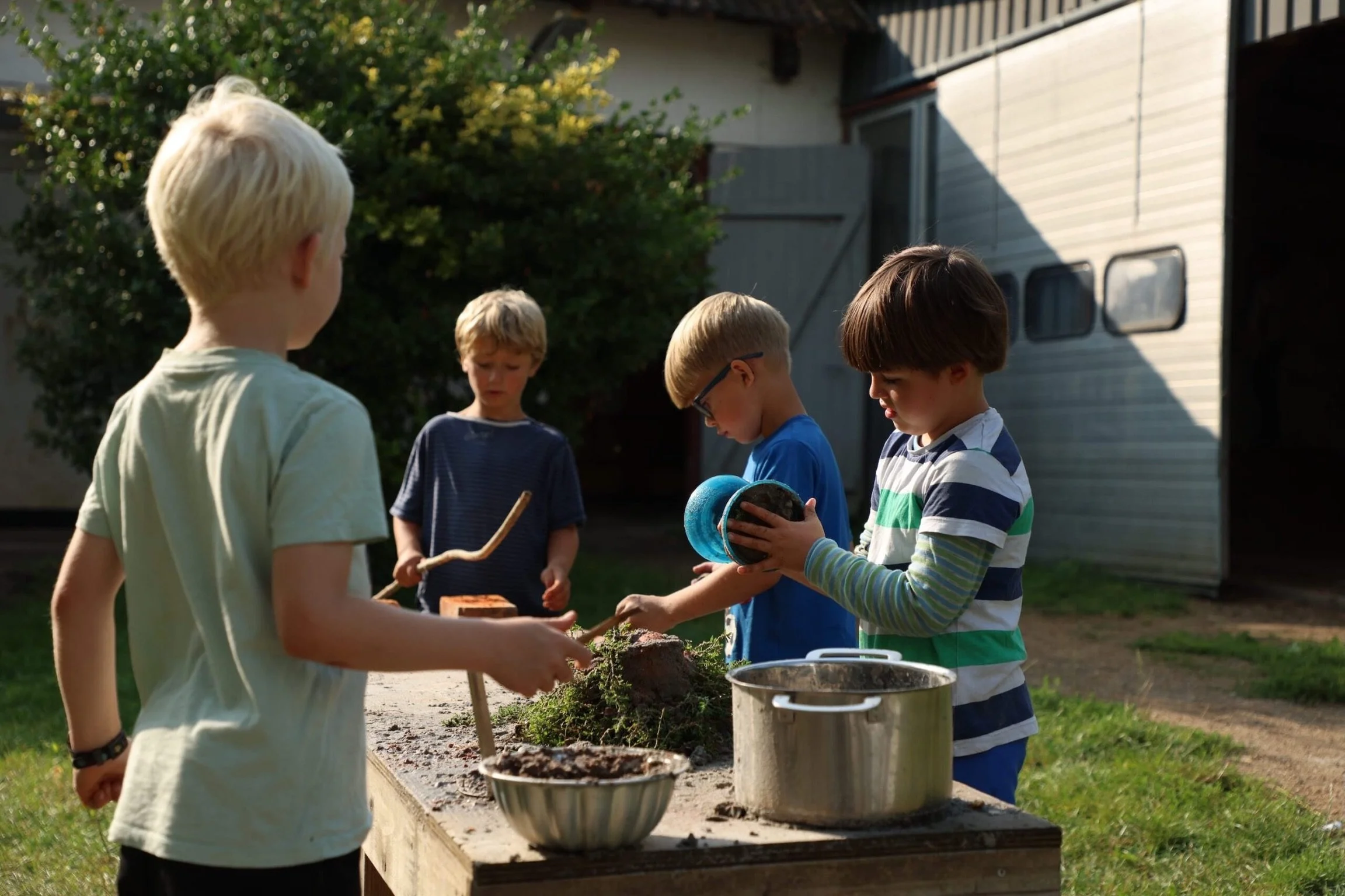 Børn leger med jord og laver aktivitet udendørs ved et træbord, med en trailer i baggrunden.