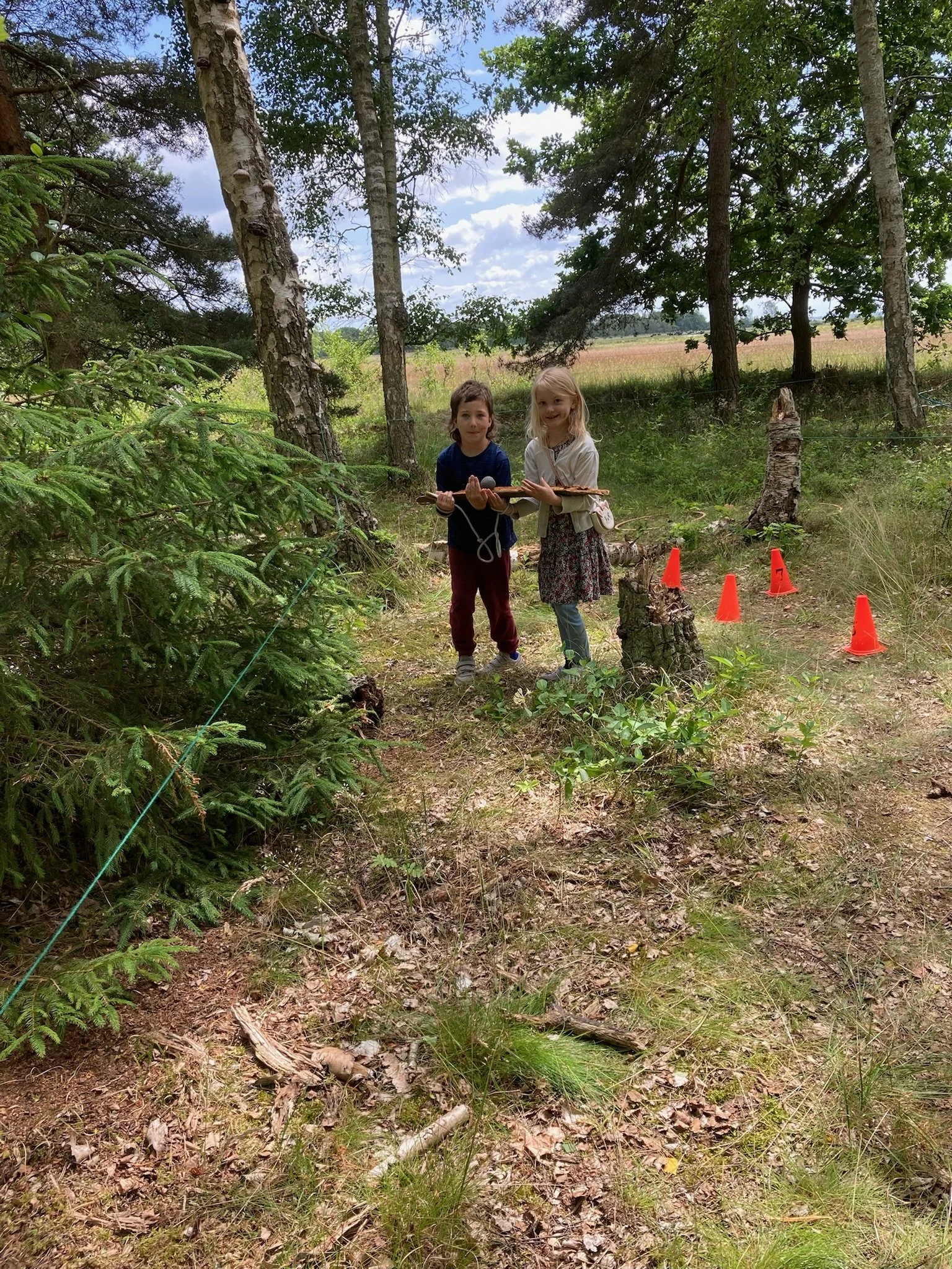 To børn i skov, holder pinde, med orange vejstrimler i baggrunden.