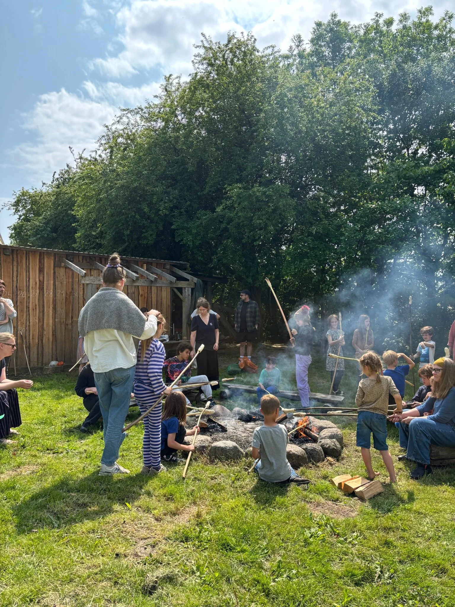 Billede af en gruppe mennesker, herunder flere børn, der laver mad over åbent bål i en have, med grønne træer og en træplads i baggrunden.