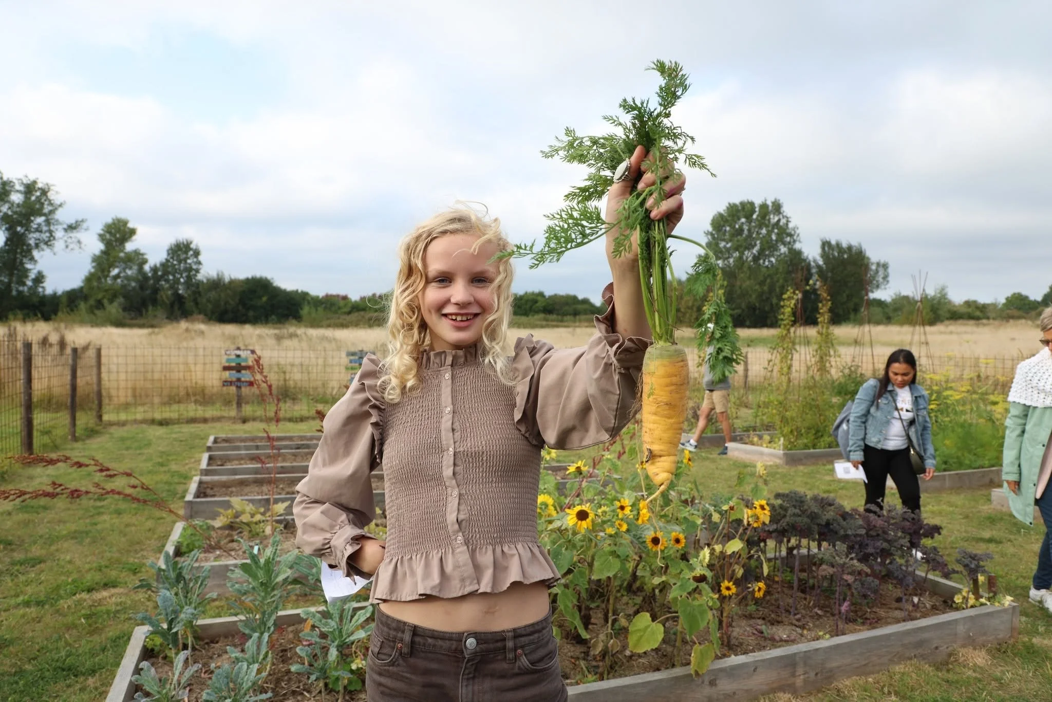 En smilende pige holder en stor gulerod øverst i haven med blomster og havearkiv i baggrunden.