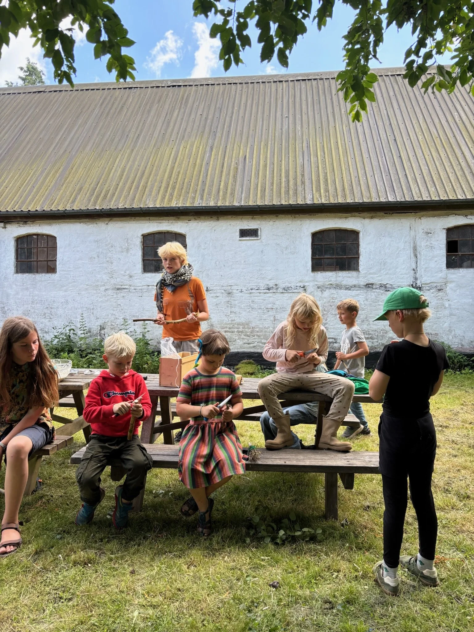 En gruppe børn sidder og står omkring et picnicbord udenfor, nogle bruger smartphones, mens en voksen står bagved. Baggrunden er en hvid bygning med små vinduer og et tag.