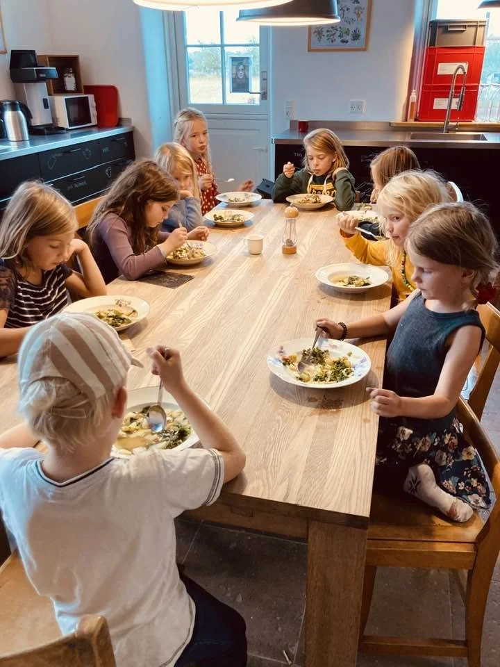 Børn sidder omkring et langt træbord og spiser. Der er ni børn, der spiser mad på tallerkener, i et lyst køkken med store vinduer og moderne apparater.