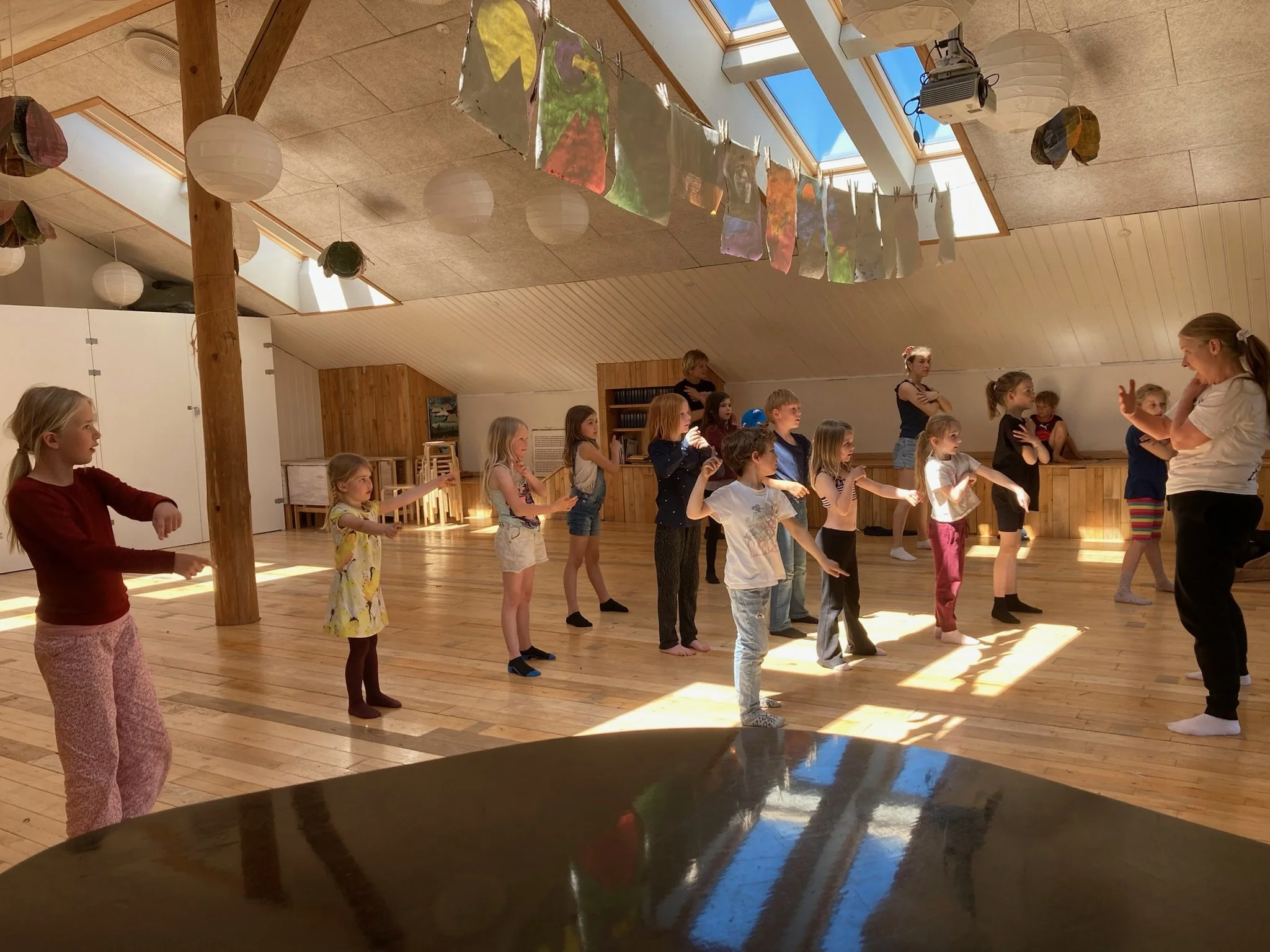 Børn deltager i en danseundervisning i en træningssal med træbjælker, lysindfald fra ovenlys, papirlamper og farverige kunstværker hængende oppe i loftet. En voksen underviser instruerer børnene.