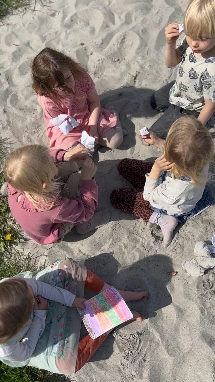 Børn sidder på sandet på stranden, der leger med papirfugle og tegner på papir.