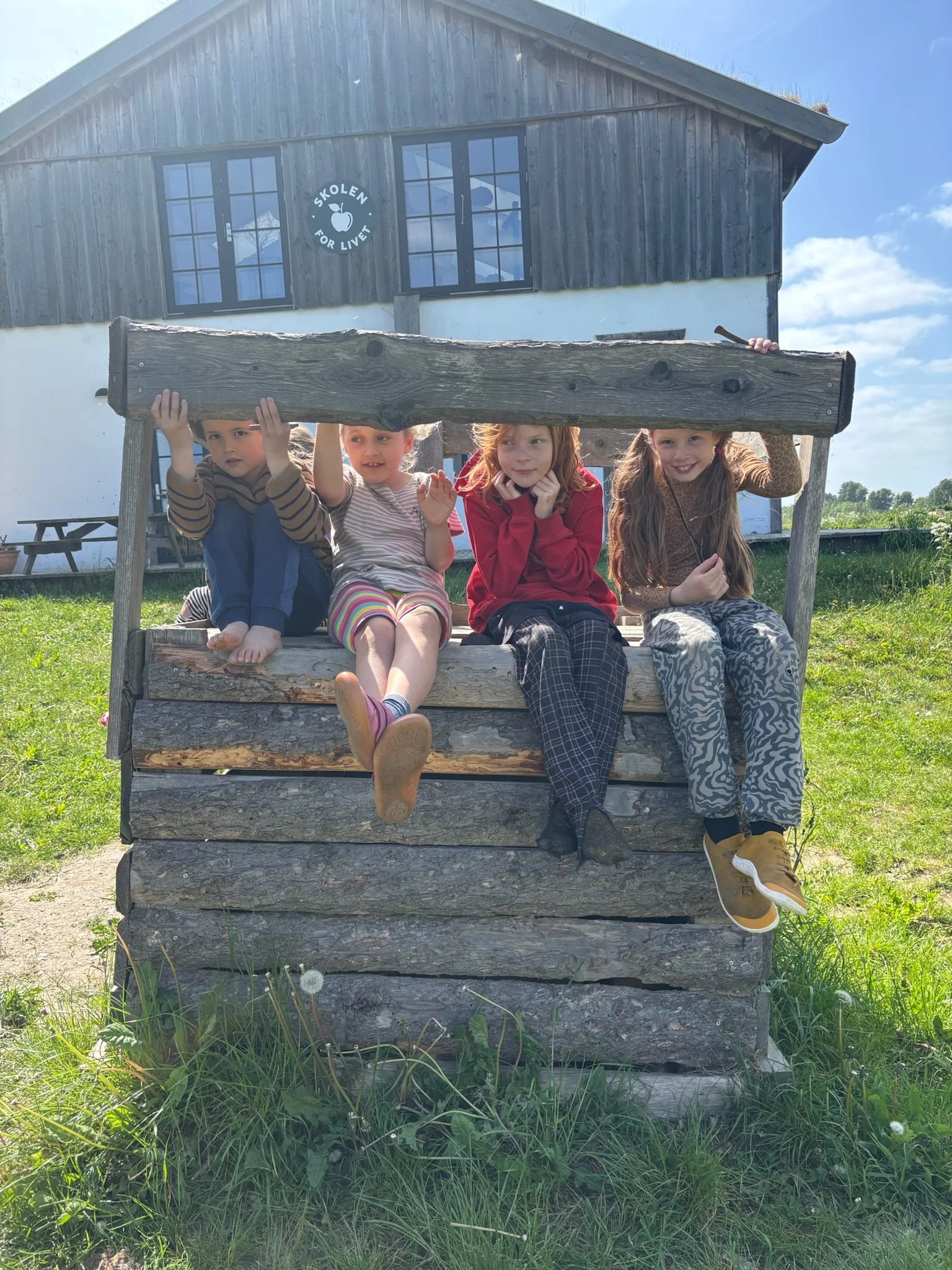 Fire børn sidder på en træbænk foran en bygning med skolen 'Skolen for Livet' skrevet på vinduet.