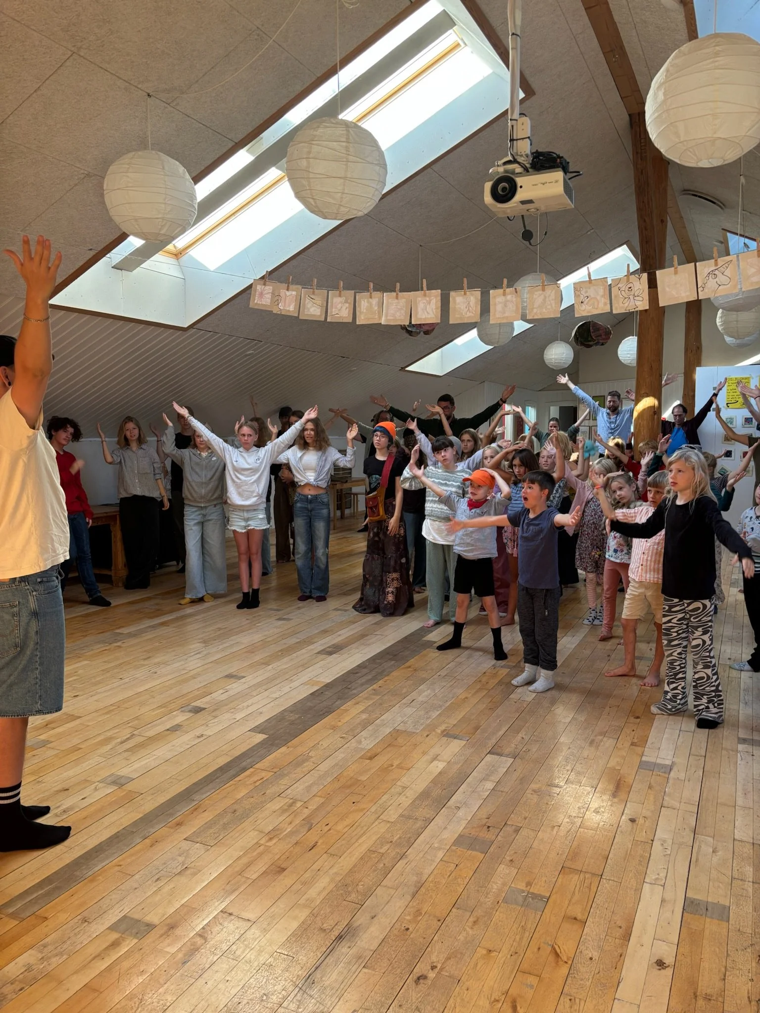 En gruppe børn og voksne, der står i en cirkel med arme hævet i et lyst rum med træloft, papirlamper og skydedøre, der vender mod en loftsdam.