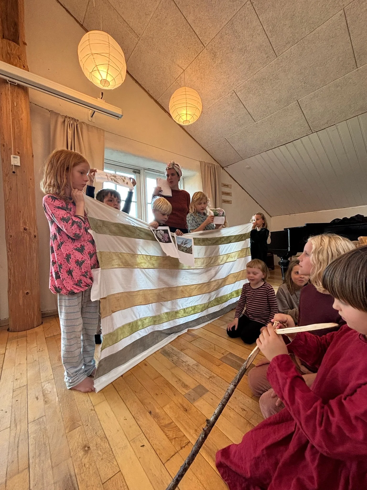 Børn holder og præsenterer en stor stribet tekstil eller banner foran en gruppe børn og en voksen i en indendørs indstilling.