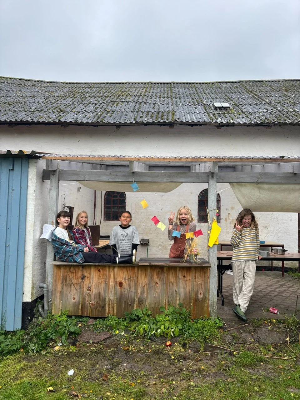 Fem børn fejrer sammen udendørs under et overdække, med papirdekorationer og smil, på en murstensebaggrund med små vinduer.