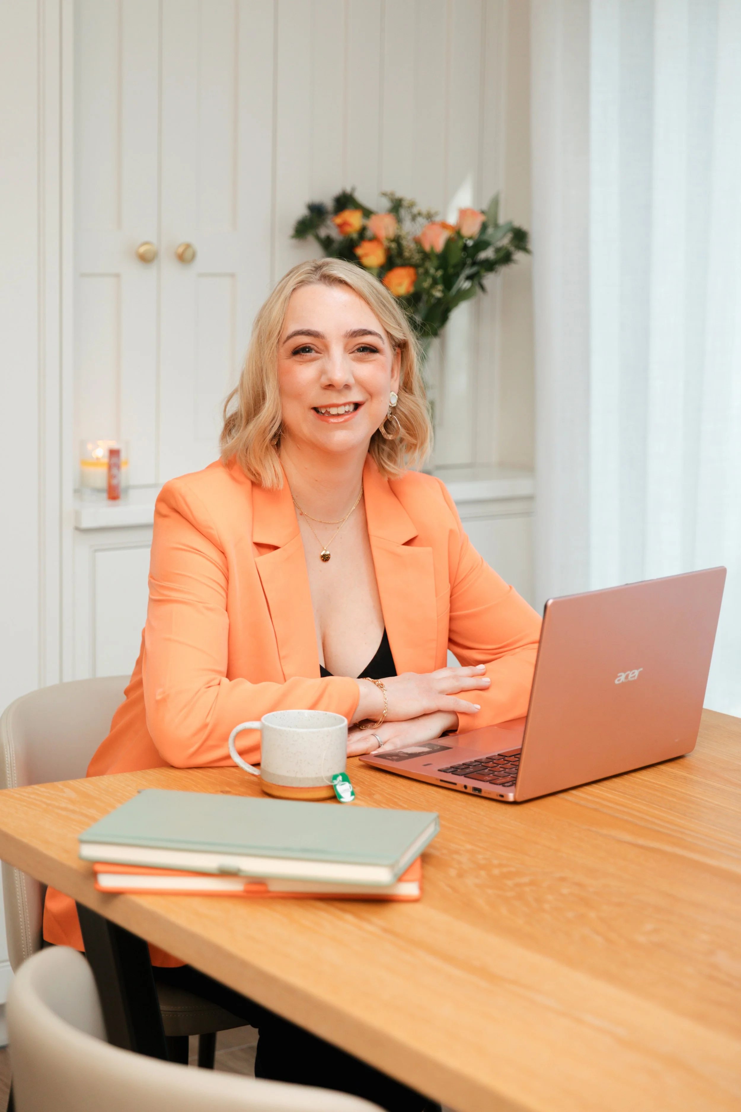 Cat, with blonde hair is sitting at a wooden table with a laptop, a mug, and some notebooks, smiling at the camera in a cozy room with white walls, a bouquet of flowers, and a window with curtains.