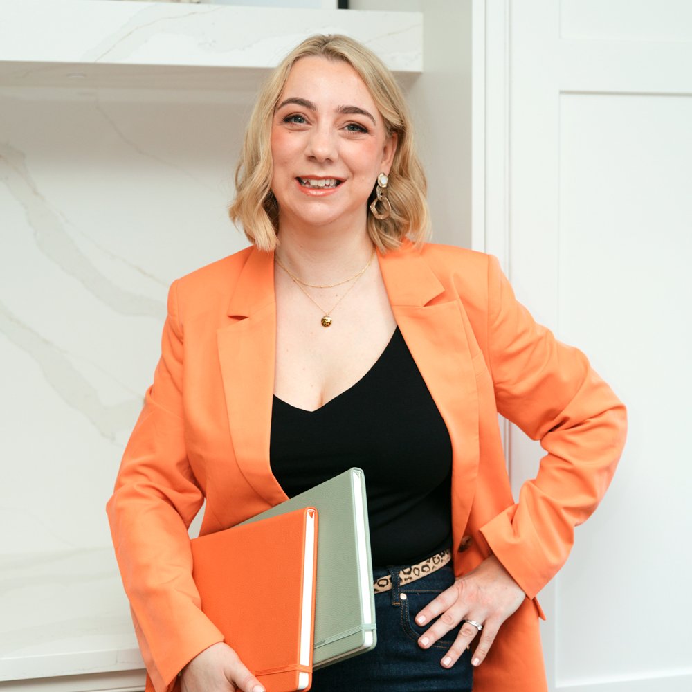 A woman with blonde hair wearing an orange blazer, black top, and holding two notebooks, standing indoors with a light-colored background.