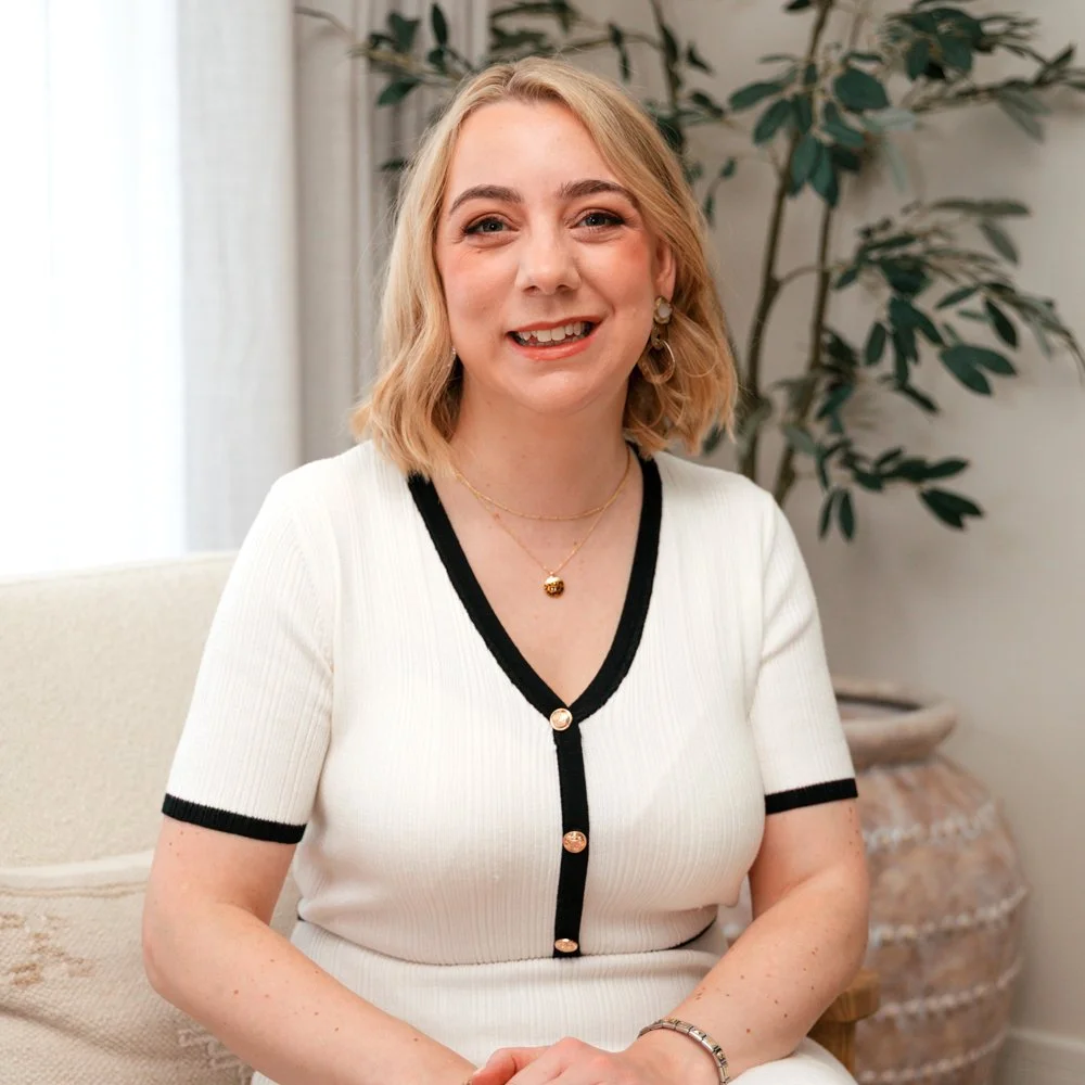 A woman with blonde wavy hair, wearing a white top with black trim, gold jewelry, and earrings, sitting on a beige chair in a room with a large plant in the background.