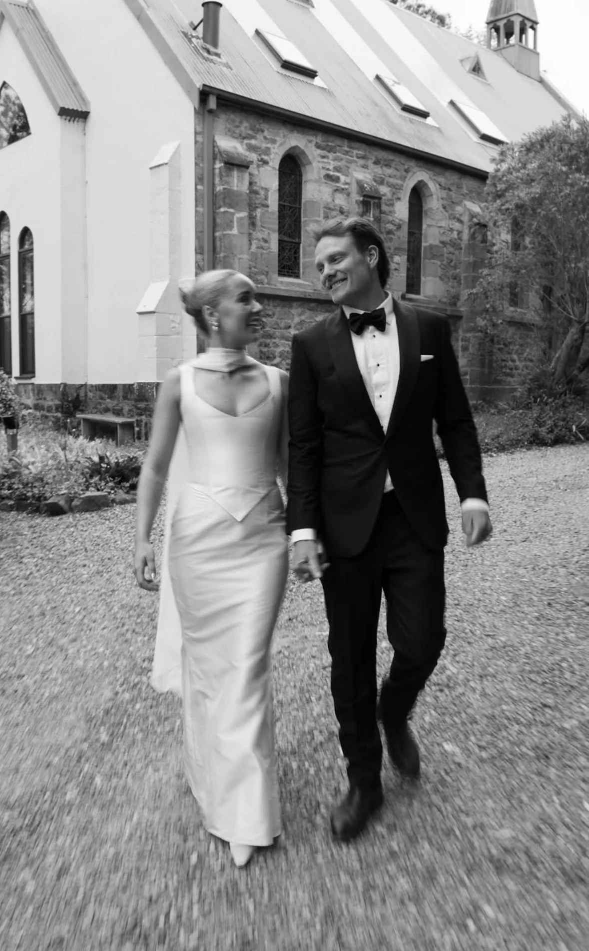 A black and white photo of a smiling couple walking outdoors in front of a stone building, dressed in formal attire. The woman is wearing a white gown, and the man is wearing a black tuxedo with a bow tie.