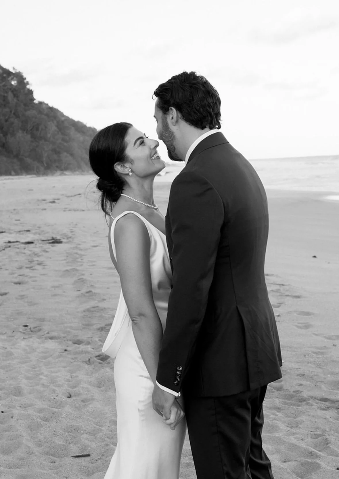 A black and white photo of a couple on the beach, standing face to face and holding hands, looking into each other's eyes.