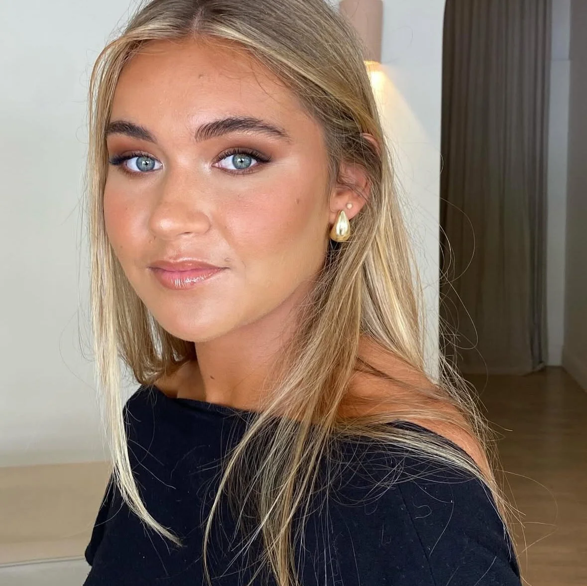 Close-up of a young woman with blonde hair, blue eyes, makeup, gold earrings, and wearing a black top, smiling softly indoors.