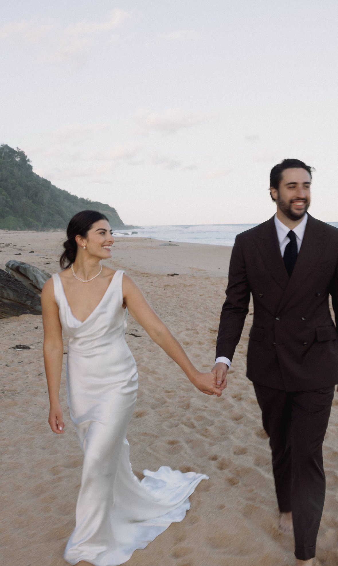 Bride and groom walking hand in hand on a beach.