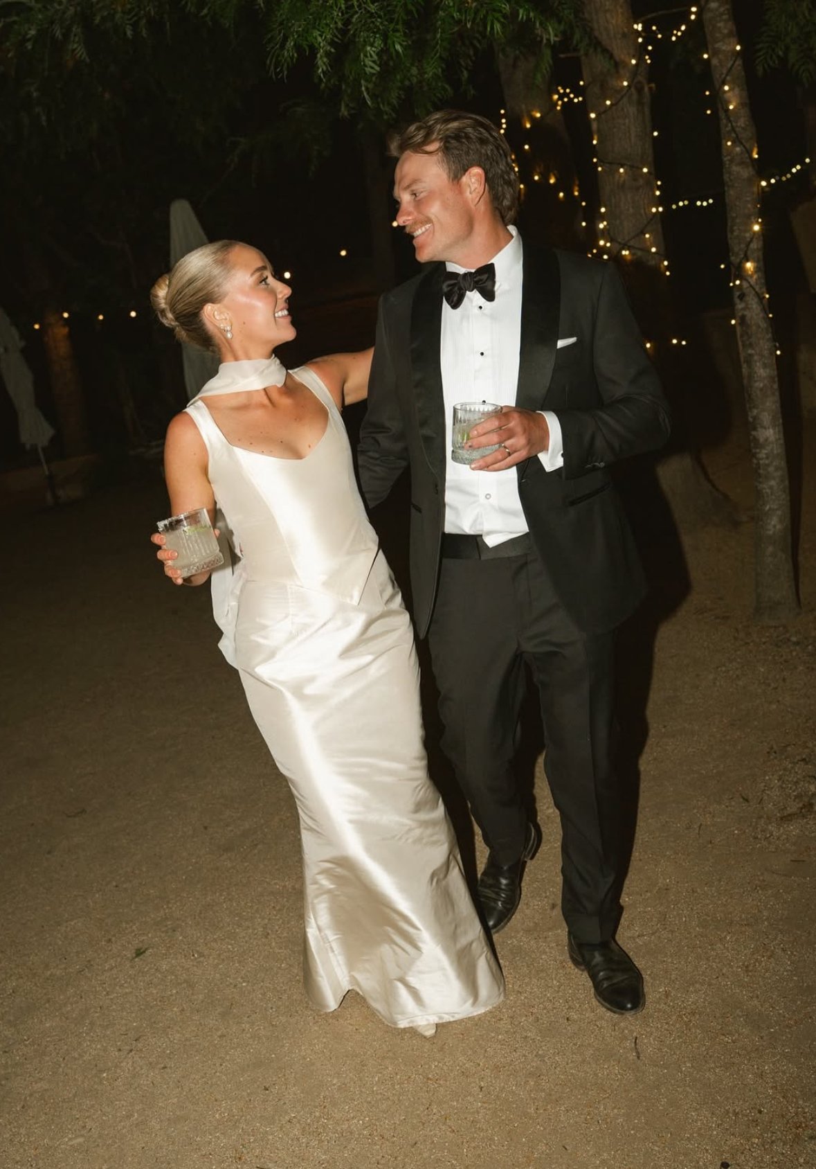 A bride and groom in wedding attire walking together at night, smiling and holding drinks, with string lights hanging in the background.