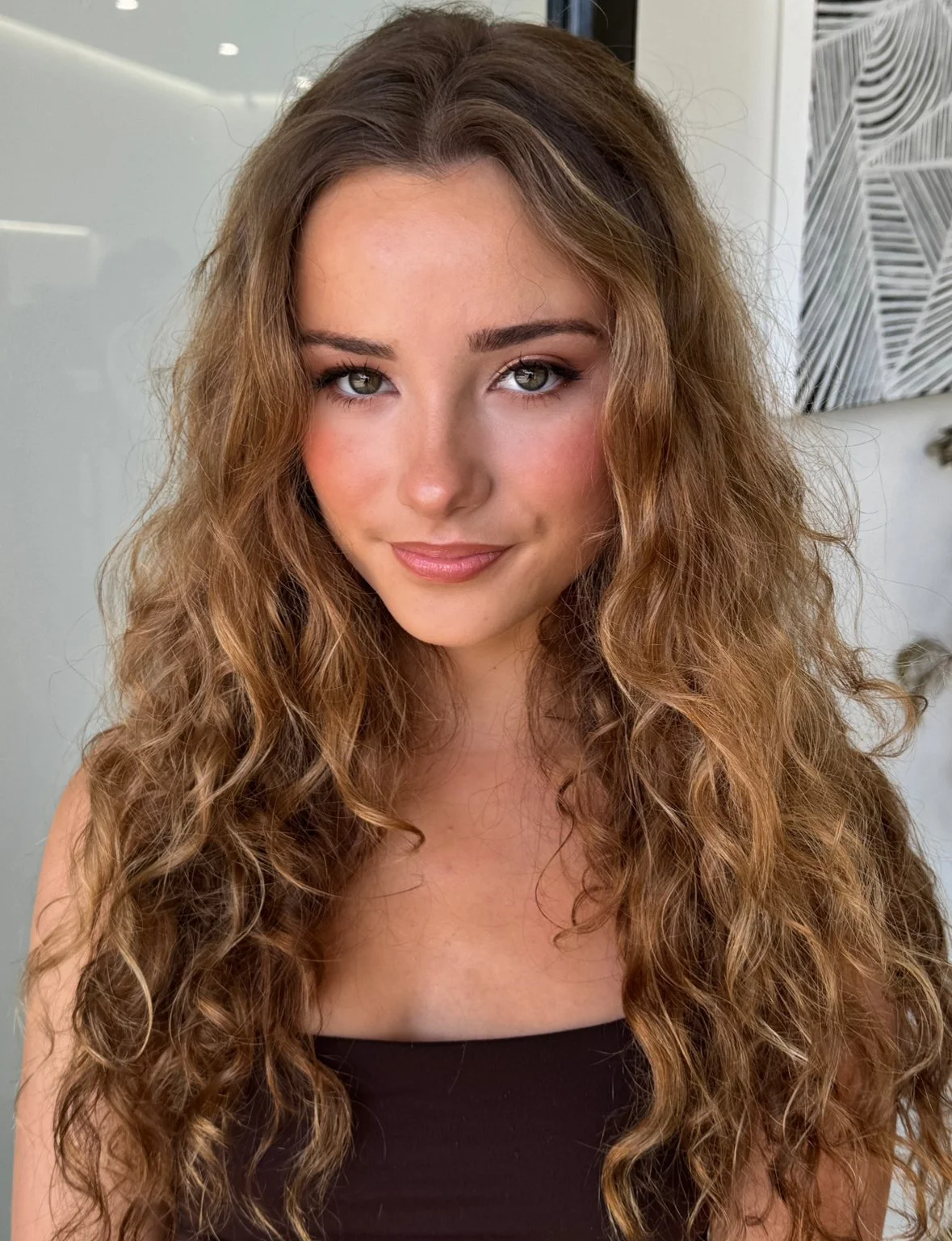 Close-up portrait of a young woman with long, curly light brown hair, fair skin, and green eyes, smiling softly at the camera. She is wearing a black top, and the background includes a white door and modern wall art.
